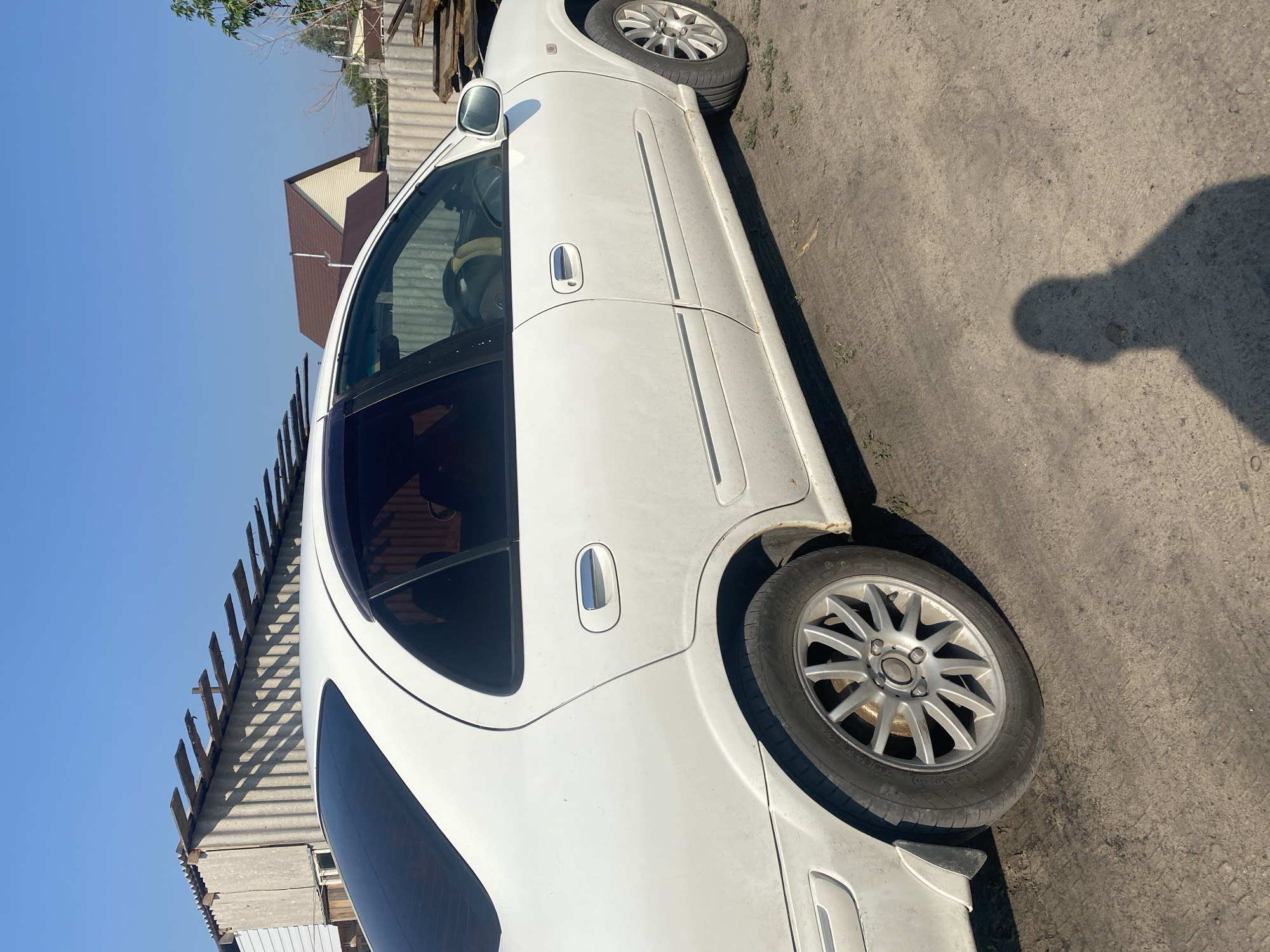 A white four-door sedan, viewed from the right rear quarter panel, is parked on a dirt surface. The car has tinted rear windows and chrome door handles. The background shows a corrugated metal fence, a wooden structure with exposed beams, and a residential building with a brown roof. The sky is clear and blue, indicating a sunny day. A shadow of a person is cast on the ground in the foreground. The location is identified as Mamontovo, Russia.