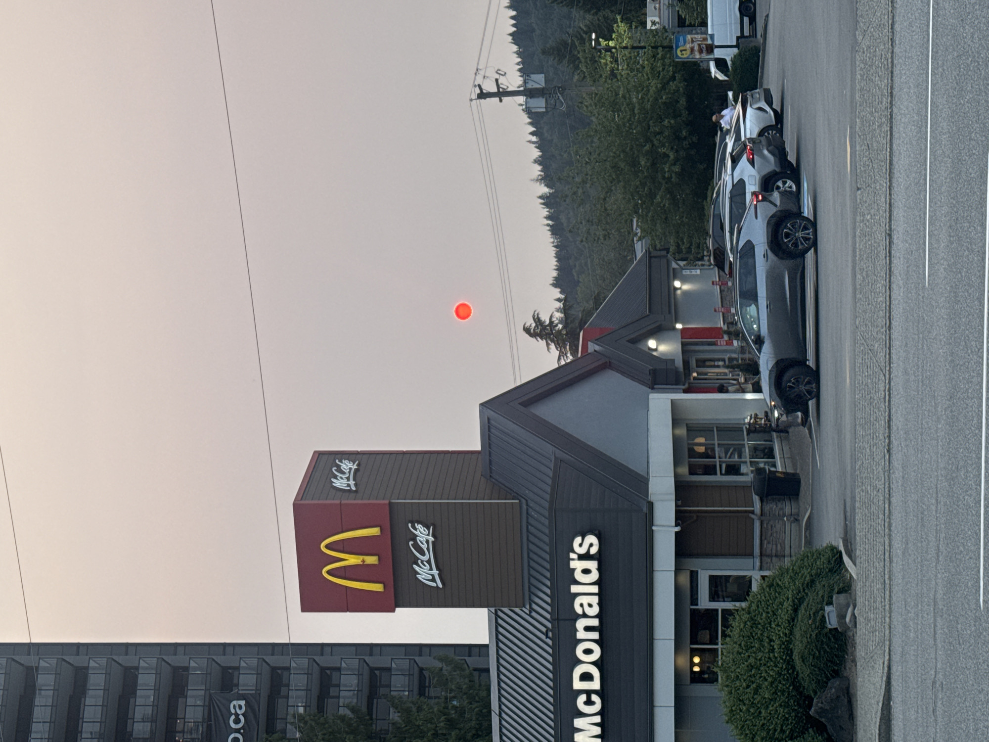 The image depicts a McDonald's restaurant in Coquitlam, Canada, under a hazy, smoke-filled sky, causing the sun to appear as a distinct red orb. A tall apartment building is visible to the left of the McDonald's sign, which prominently features the iconic golden arches and "McCafé" branding. Several vehicles are parked in the restaurant's lot, including a grey SUV in the foreground. Trees and power lines are present in the background, with a dense forest line visible on the horizon. The overall atmosphere is subdued due to the smoky conditions.