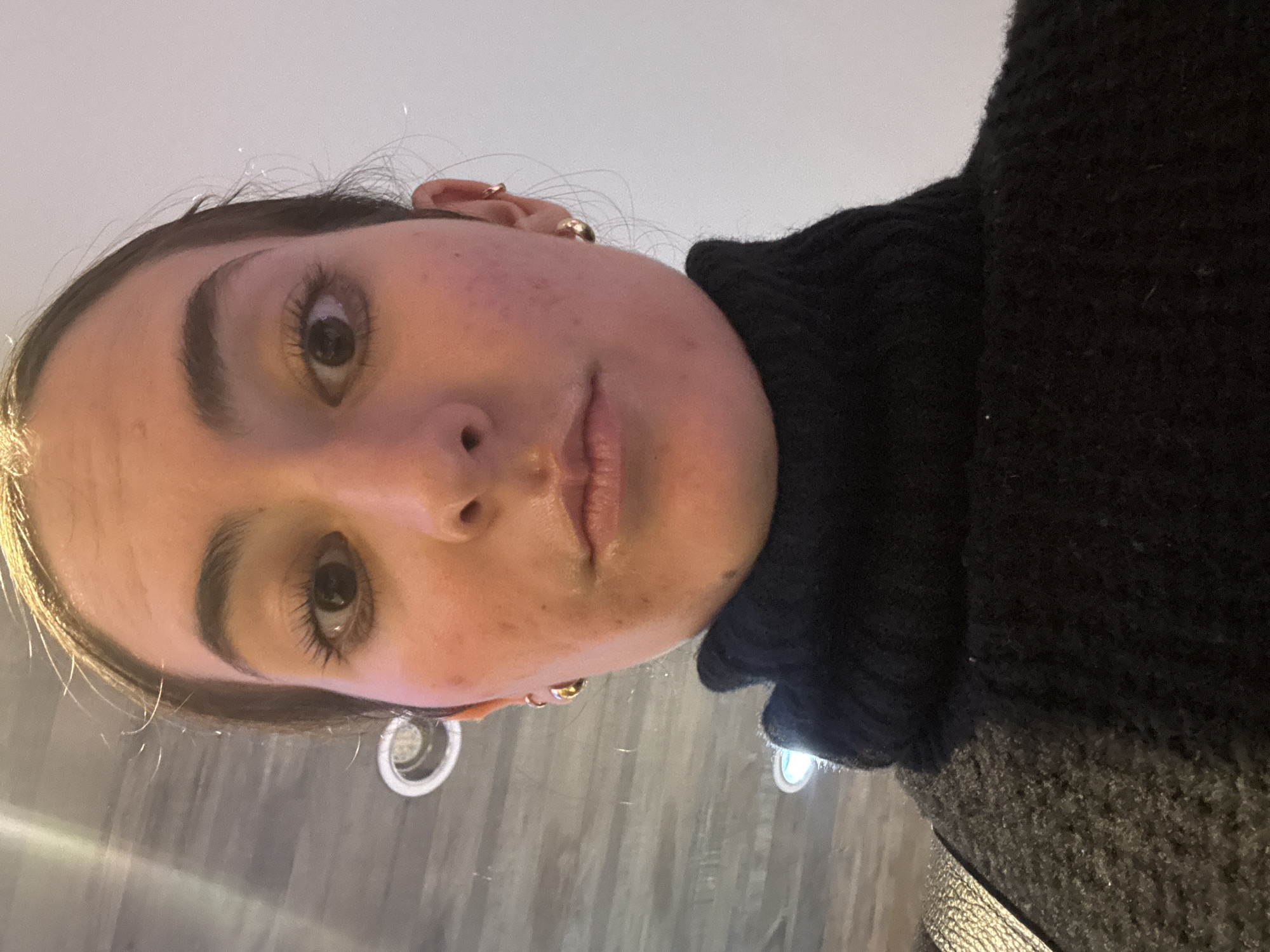A close-up, low-angle selfie of a fair-skinned person with brown eyes and dark eyebrows, wearing a black turtleneck sweater. Gold earrings are visible. The background is nondescript, possibly an interior with lighting fixtures. There's no indication of the location being Puebla, Mexico, within the image itself.