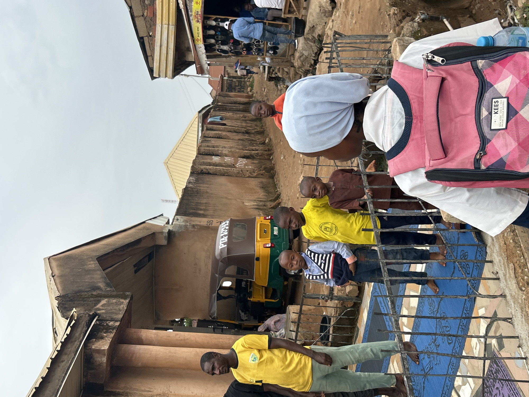 The image captures a vibrant street scene in what appears to be an urban or semi-urban area of Jos, Nigeria, under an overcast sky. The ground consists of dirt and some tiled areas, with concrete buildings and corrugated metal roofs forming the backdrop.
In the foreground, a group of people is visible. To the left, a man in a yellow t-shirt and light green pants stands, looking towards the viewer. To his right, a blue patterned mat is spread on the ground behind a dark metal barrier or railing. Several barefoot children are gathered on or near this mat; one wears a yellow t-shirt, another a striped long-sleeve shirt with jeans, and a third a brown tunic. Behind them, partially visible, a person in a white top with a light blue head covering, and another child in an orange top, are also present.
A prominent feature in the midground is a yellow and green motorized tricycle, often called a "keke napep," parked between buildings. Visible text on the tricycle includes "PTA-10345" on the side and "PTA-1" on the front. In the background, there's a shop displaying merchandise, likely shoes, and a man in a light blue shirt is standing near it.
To the right side of the frame, a pink and dark blue plaid backpack with a zipper is resting, along with a clear plastic water bottle. Text on the backpack clearly reads "KEES," "NEW YORK SINCE 1903," and "MAJOR LEAGUE BASEBALL." Another small sign with "FB NOW" is visible on a building in the background. The scene depicts a slice of daily life with people gathering outdoors.