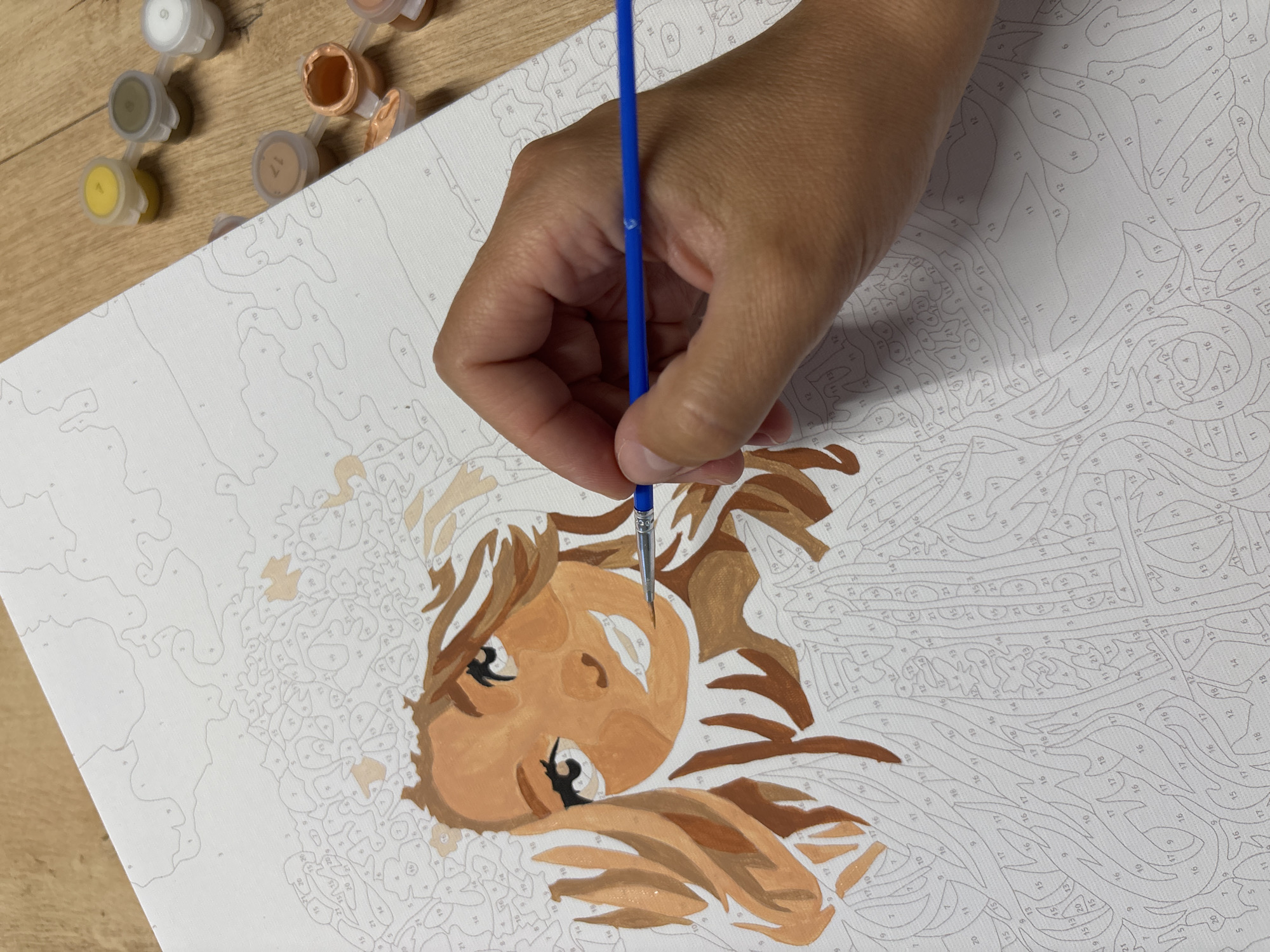 The image depicts a person's hand holding a paintbrush and applying paint to a "paint by numbers" canvas. The canvas features an unfinished portrait of a woman with brown hair and a light complexion. Several small containers of paint are visible in the background, along with a wooden surface, likely a table. The scene is a close-up, focusing on the painting process. There is no information available to determine the location of this image within Chaikovsky, Russia.