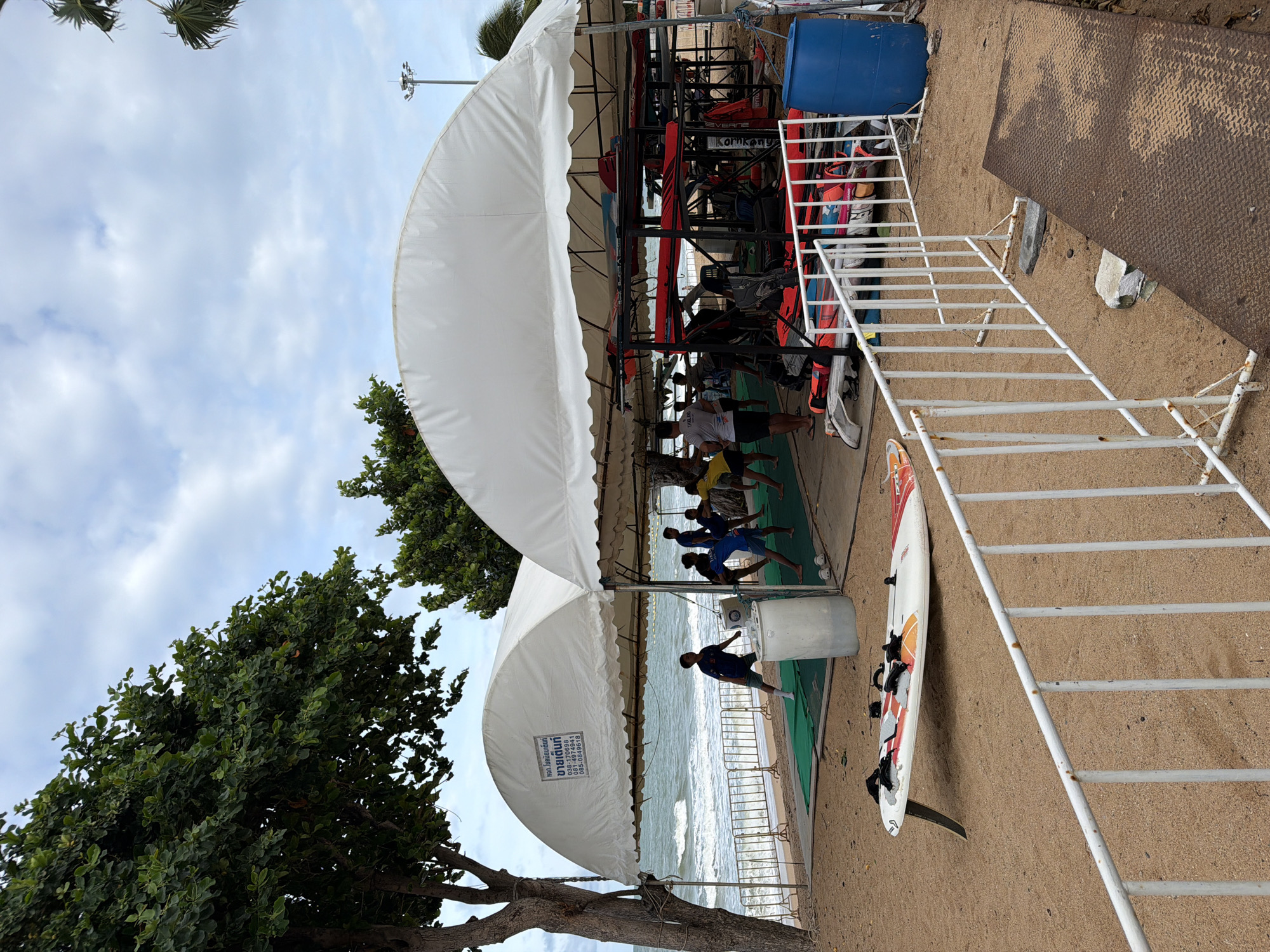 This image depicts a coastal scene in Pattaya City, Thailand, featuring a watersports or training facility set up on a sandy beach. The foreground presents light brown sand. A white metal barrier, showing signs of rust at its base, extends horizontally across the lower portion of the frame. To the left of this barrier, a white and orange windsurfing board, equipped with black foot straps and a fin, lies on the sand. To the right, a rusted metal tread plate is partially visible on the sand.
Dominating the midground are two large, white, arched canvas tents. The tent on the left features Thai script on its front, which includes "ขายเต็นท์" (Sell Tent) and contact phone numbers (038-170688, 081-3532881, 085-0849618). Under this tent, several individuals, primarily young males, are engaged in a group exercise or training session on green mats laid over the sand. One boy in a blue shirt stands near a white plastic barrel. Others are positioned further back, performing various movements.
The tent on the right is larger and appears to function as storage for watersports equipment. Numerous surfboards or windsurfing boards are stacked on black metal racks. Red, blue, and other colored sails or board components are visible alongside various bags, life vests, and other gear arranged on the sand beneath the racks. A blue plastic barrel is situated at the far right edge of this tent area. One person wearing a white t-shirt with "THAILAND" on the back is visible under this tent.
In the background, beyond the training area, another metal barrier separates the sandy beach from the ocean. Waves are breaking on the shore, and the sea extends to the horizon under a partly cloudy sky. A large tree with dense green foliage frames the left side of the image, and another tree is partially visible behind the right tent. A tall utility pole with multiple light fixtures stands in the distance on the right, and the fronds of a palm tree are visible in the upper right corner.