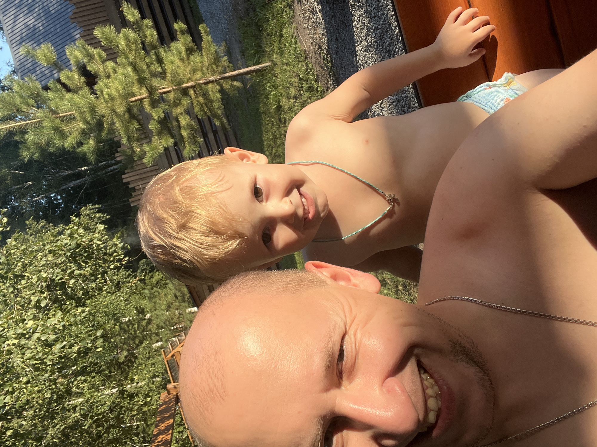 The image is a selfie taken outdoors on a sunny day. The foreground features the face of a man, who appears to be smiling and looking at the camera. He is bald and wearing a silver chain. Behind him, a young child with blonde hair is also looking at the camera, smiling and wearing a light blue necklace. The child is wearing a diaper with a subtle pattern. The background shows greenery, trees, and what appears to be a wooden structure, possibly a fence or part of a building. The overall setting suggests a relaxed, casual outdoor environment. The lighting indicates it is likely daytime, with the sun casting shadows and illuminating the subjects. The location is specified as Omsk, Russia.