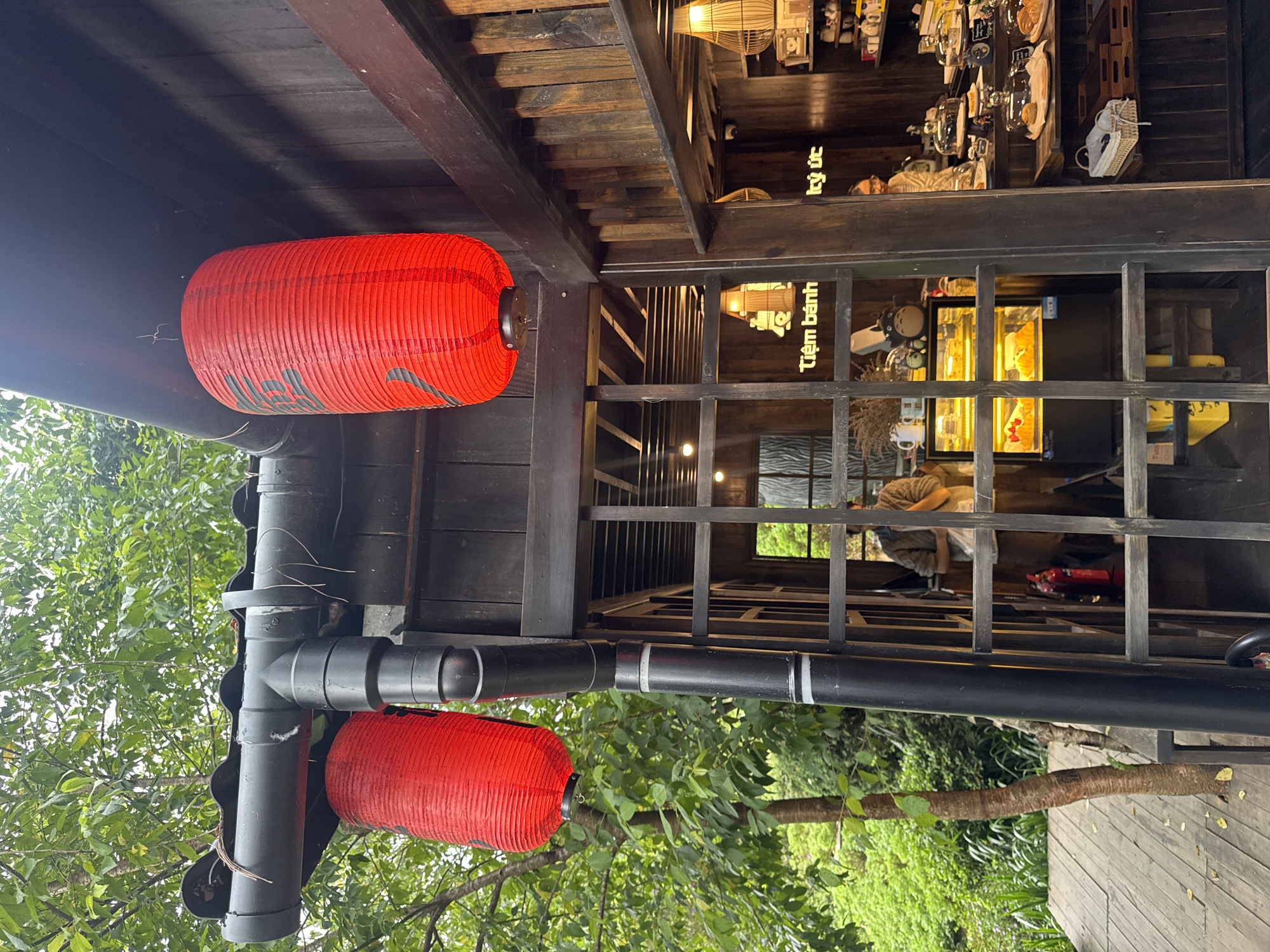 This image shows the exterior of a rustic wooden building, likely a cafe or bakery, with an Asian-inspired architectural style. Two large, red paper lanterns with black text hang from the eaves of the roof. A black downspout runs vertically along the side of the building, adjacent to a wooden lattice structure. Through the lattice, the interior of the building is visible, revealing display cases filled with pastries, shelves stocked with various items, and a person standing behind a counter. The building is surrounded by lush green foliage and trees, suggesting a natural or park-like setting. The signage inside the building, "Tiệm bánh," indicates it is a cake shop. The presence of a fire extinguisher on the right side of the lattice suggests a safety measure. The overall impression is one of a charming, traditional establishment nestled within a verdant environment. While the image does not explicitly show the city of Cam Ly - Đà Lạt, Vietnam, the architectural style and vegetation are consistent with the region's aesthetic.