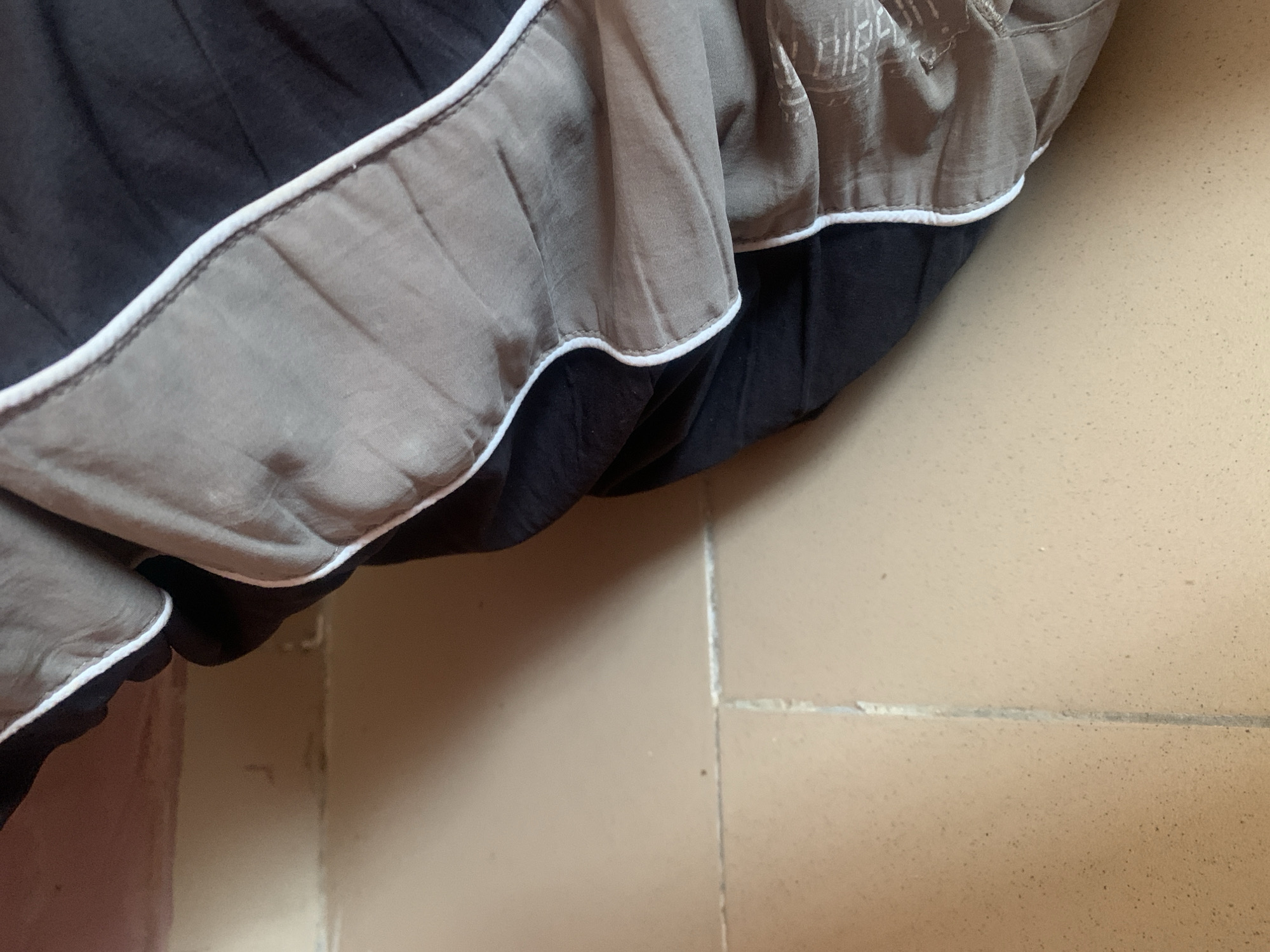 The image shows a close-up view of a folded comforter or duvet, primarily in black and grey tones, with a white trim. The fabric appears to be laid on a tiled floor. The tiles are a light brown or beige color, and visible grout lines suggest they are laid in a grid pattern. The lighting suggests an indoor environment, possibly a bedroom. The scene is devoid of people or any other living beings. The specific location in the city of Jauro Yinu, Nigeria cannot be determined from the visual information provided in the image.
