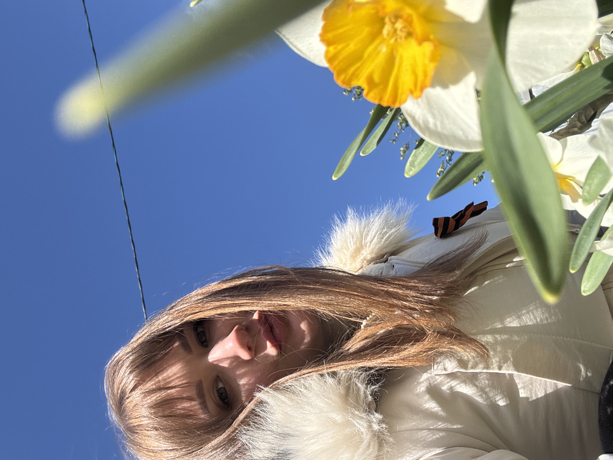 A low-angle close-up captures a woman with light brown hair, partially obscuring her face, under a bright blue sky. A white and yellow daffodil flower is prominently positioned in the upper right of the frame. A thin wire extends across the upper left portion of the sky. The woman is wearing a light-colored jacket with a fur collar and an orange and black ribbon is visible attached to the jacket, near the top edge of the frame. Based on the presence of the St. George's ribbon, and given the prompt's information about Chaikovsky, Russia, the scene likely relates to commemorations of Victory Day in Russia.