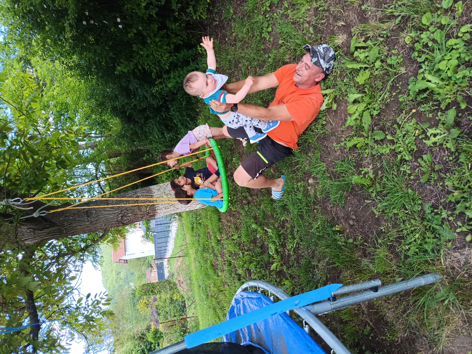 The scene depicts an outdoor recreational area in Kovachevtsi, Bulgaria, characterized by natural elements and human activity. An adult male lies supine on a patch of grass and dirt in the foreground, wearing an orange short-sleeved shirt, dark shorts with a light stripe, blue and white striped slide sandals, and a camouflage-patterned bandana. He is actively holding a baby aloft with both arms extended upwards. The baby, dressed in a light blue and red striped t-shirt, patterned grey pants, blue shoes, and a white bib, has outstretched arms and appears engaged in play.
In the mid-ground, to the left of the central figures, a large tree with green foliage anchors a green circular swing seat suspended by yellow ropes. Three children are present near the swing. One boy sits on the swing, wearing a blue t-shirt and dark shorts, looking towards the right. Another child, a girl in a light purple t-shirt and patterned shorts, stands nearby, partially obscured. A third child, a boy in a dark t-shirt, is partially visible to the left of the swing.
To the far left, a portion of a trampoline with a blue mat and metal frame is visible. The ground consists of mixed grass, weeds, and exposed soil. The background features dense green foliage, several trees, and distant green hills. A white house with a red-tiled roof is discernible in the mid-background, along with a modern dark fence and wire fencing delineating properties. The sky, visible through the tree canopy, appears overcast.