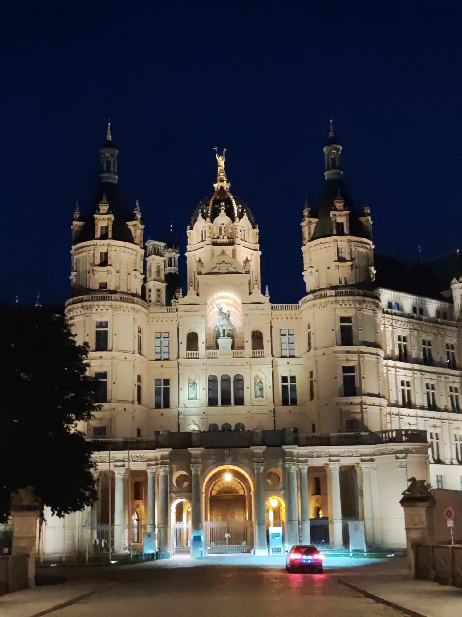 The image shows Schwerin Castle in Schwerin, Germany, illuminated at night. The castle is a grand, ornate structure with multiple towers and a large central dome, all lit with warm yellow light. A dark tree is visible on the left side of the frame. In front of the castle, a dark-colored car with its brake lights illuminated is driving away from the viewer on a paved road. The sky is a deep, dark blue, indicating nighttime.