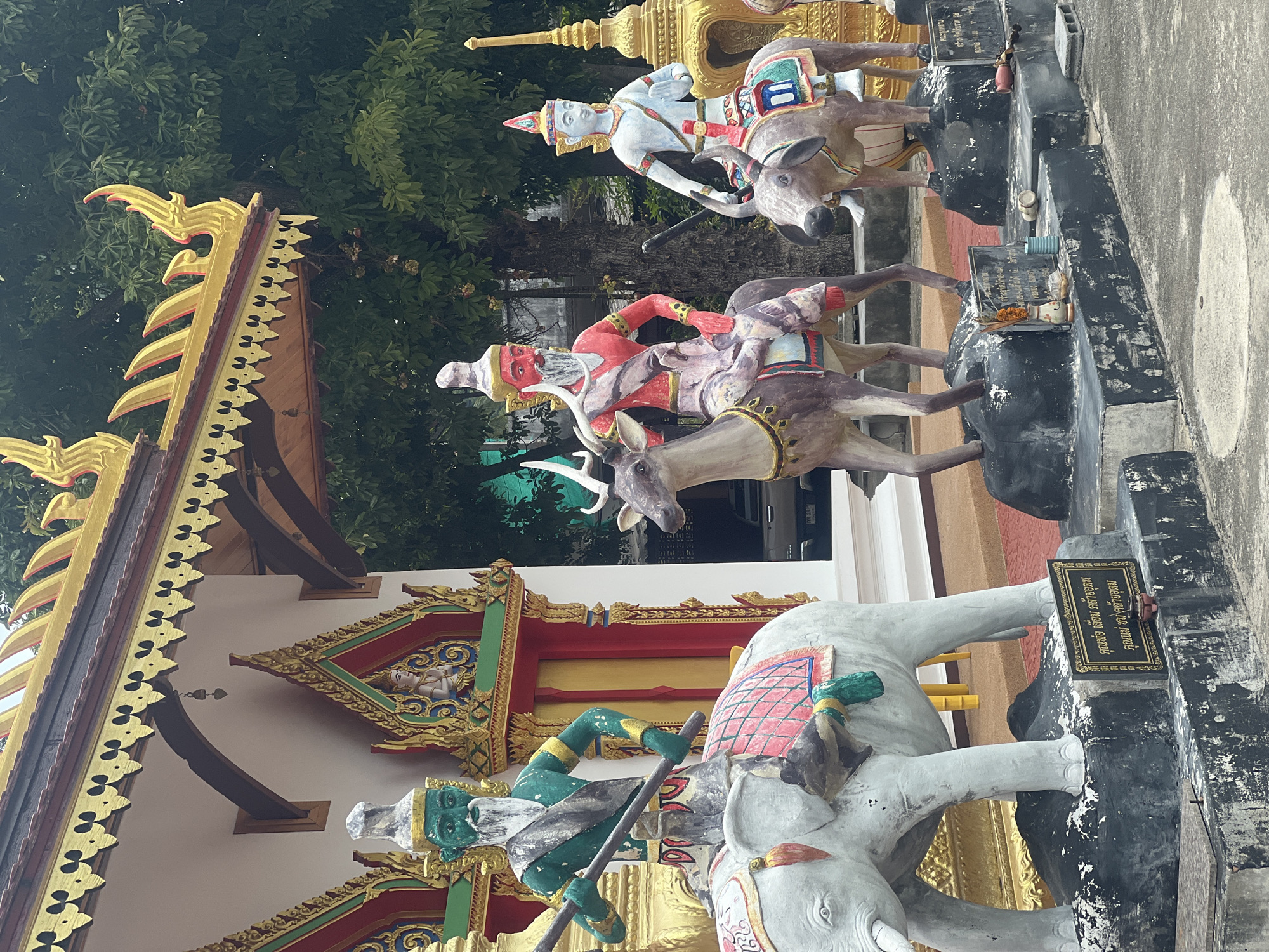 In an outdoor setting in front of a building with a red and gold ornate roof, three statues are displayed. On the left, a large white elephant statue stands with a green-skinned figure riding on its back. The figure wears a white headdress, red sash, and holds a long staff. To the right of the elephant, a brown deer statue is depicted with a red-skinned figure as its rider. This figure also has a white beard and wears a white headdress. Further to the right, a brown bull-like creature statue is shown with a white-skinned figure riding it, wearing a conical headdress. The figures are adorned in colorful clothing and headdresses. Behind the statues, the building exhibits intricate gold trim and decorative elements. Trees and foliage are visible in the background, suggesting a possibly tropical environment. A small plaque with Thai writing is placed at the base of the elephant statue. Several other small offerings and objects are scattered around the bases of the statues.