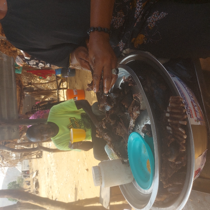A person arranges cooked meat on a large metal platter alongside ribs. The platter holds a small turquoise plate with spice and a white container. A man in a green shirt holding a yellow cup sits in the background. The setting is outdoors, near a building with visible wooden beams.