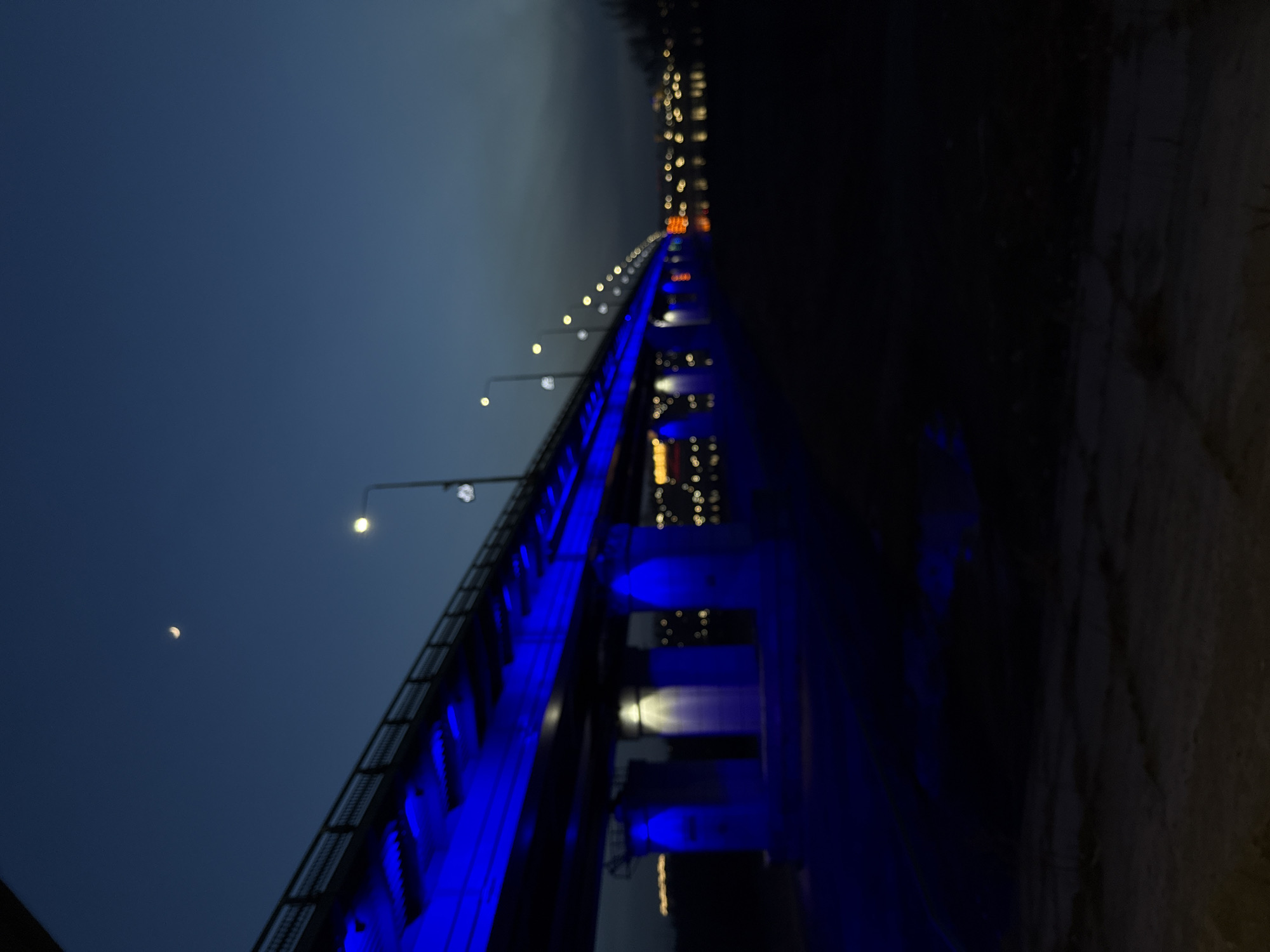 The image captures a nighttime or late dusk scene of a long bridge illuminated by vibrant blue lights. The perspective is from the side of the bridge, looking along its length as it recedes into the distance.
The central subject is the bridge, which has a prominent railing on one side, lined with conventional streetlights emitting yellow-white light. The most striking feature is the intense blue light emanating from the underside of the bridge deck and illuminating its support pillars or structure. This blue illumination creates a strong visual contrast against the dark blue sky and the surrounding darkness.
In the upper-left quadrant of the dark sky, a clear, white crescent moon is visible. In the distant background, beyond the illuminated span of the bridge, numerous blurred points of light suggest the presence of a cityscape or distant buildings. The immediate foreground on the right side of the image is very dark, showing only vague textures that could be ground or a bank.
The time of day is clearly night or deep twilight, characterized by the dark sky and the reliance on artificial lighting. The sky appears clear, as indicated by the visibility of the moon. No people or specific activities are visible. The overall mood is tranquil and atmospheric, with the blue lights giving the bridge a distinctive and artistic appearance. There is no visible text in the image.