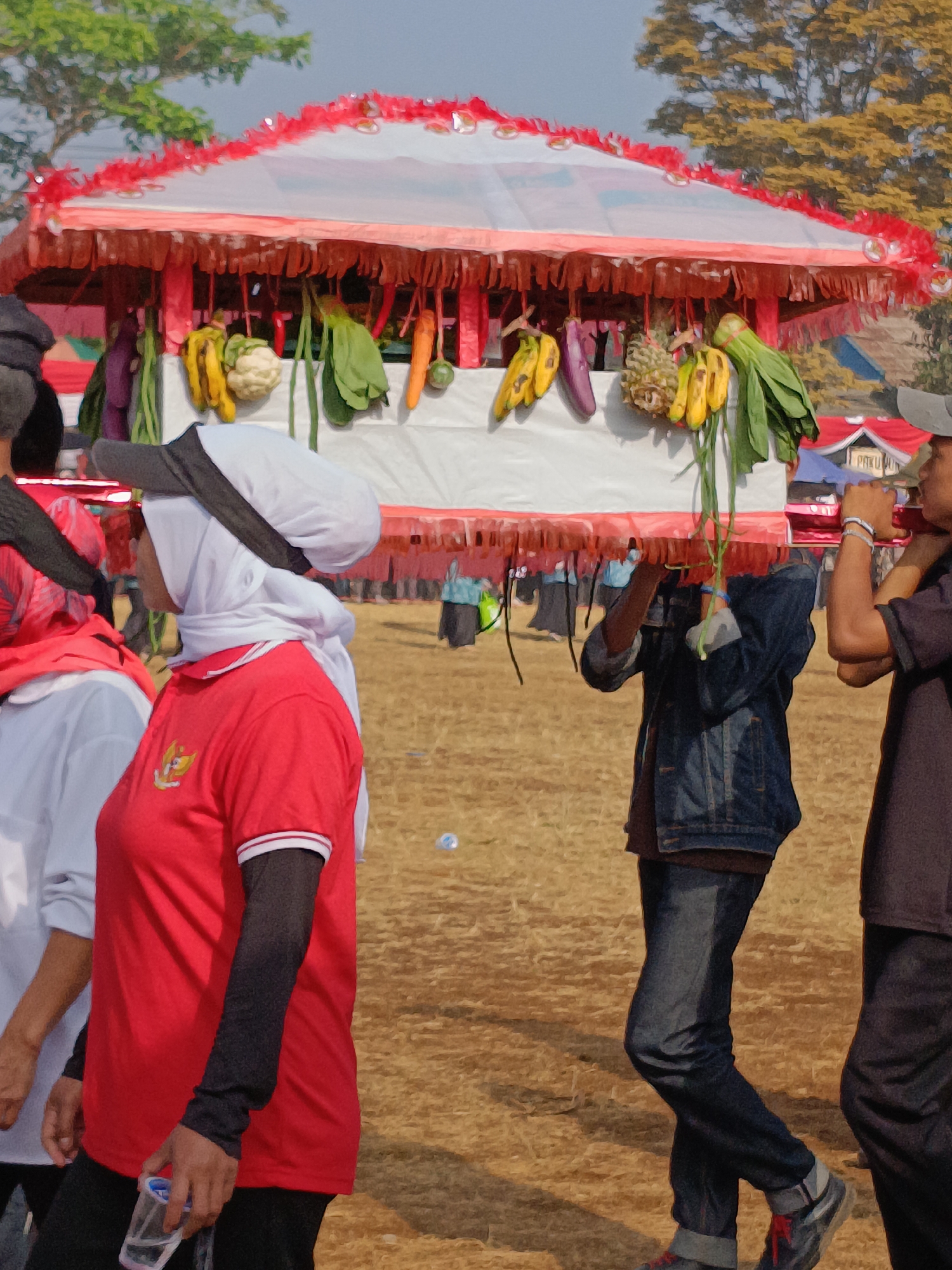 A procession is underway in the city of Cibodas, Indonesia. In the foreground, a woman wearing a red shirt and a white hijab walks to the left, holding a plastic cup. To her left, another person in a white shirt and red headscarf is partially visible. Behind them, several individuals are carrying a decorated structure suspended from poles, adorned with fruits and vegetables like bananas, eggplants, cauliflower, and carrots. This structure appears to be a canopy or a float. The ground is covered in dry, light brown soil. In the background, trees with green and yellow foliage are present, along with glimpses of buildings and other people participating in the event. The sky is a pale blue. The overall scene suggests a cultural or religious festival.