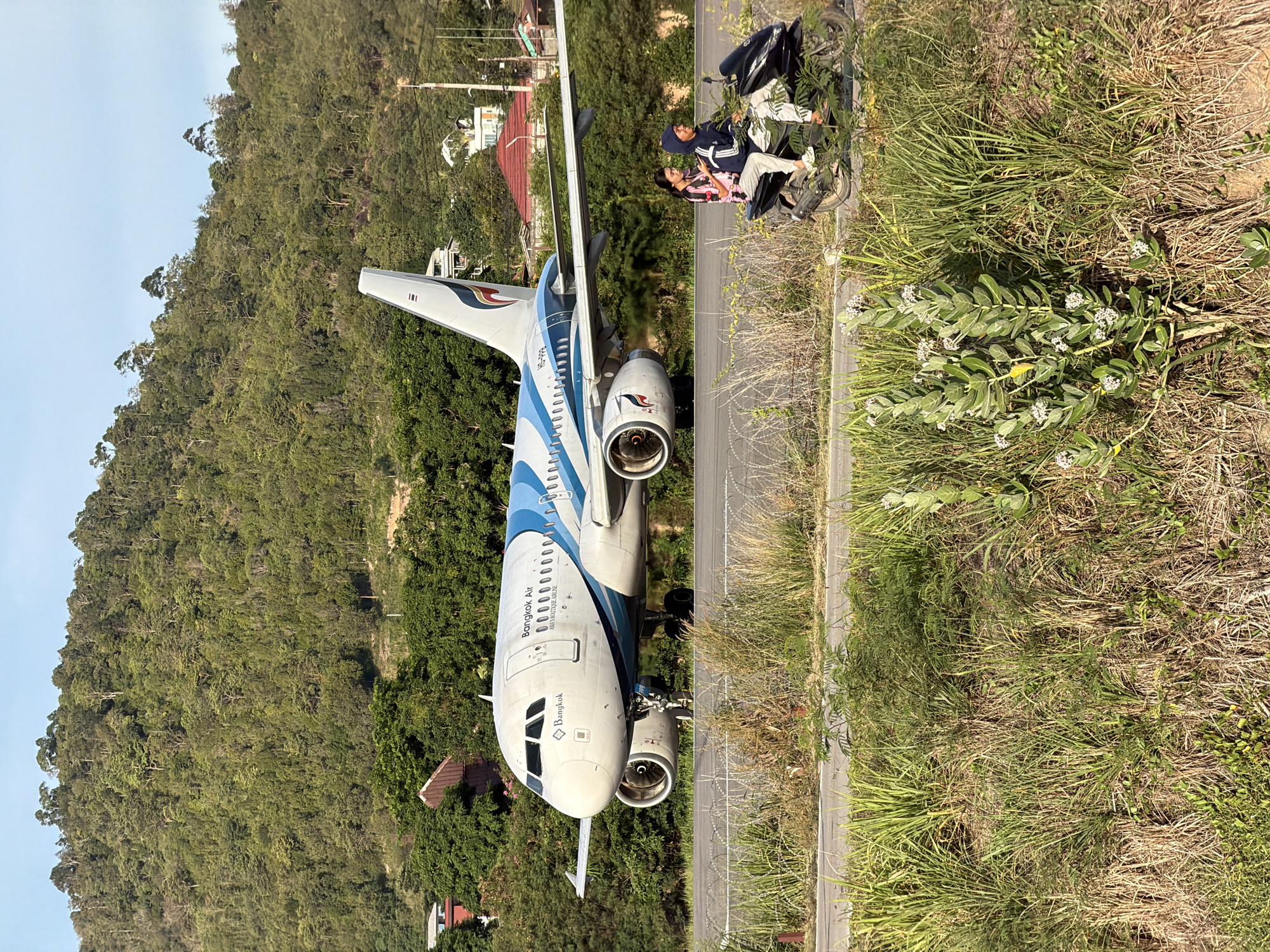 A wide shot captures an airplane on a runway with a lush, green, forested hillside in the background. The airplane, identified as Bangkok Air, is primarily white with blue accents and has "Bangkok Air" written on its fuselage. In the foreground, two individuals are riding a black scooter. The male rider wears a hooded jacket, and the female passenger wears a patterned shirt. They are positioned on a paved surface adjacent to a grassy area. The environment appears to be a tropical or sub-tropical region, consistent with Baan Bo Phut, Thailand, given the vegetation and architectural styles of the distant buildings. The lighting suggests it is daytime, with the sun casting shadows.