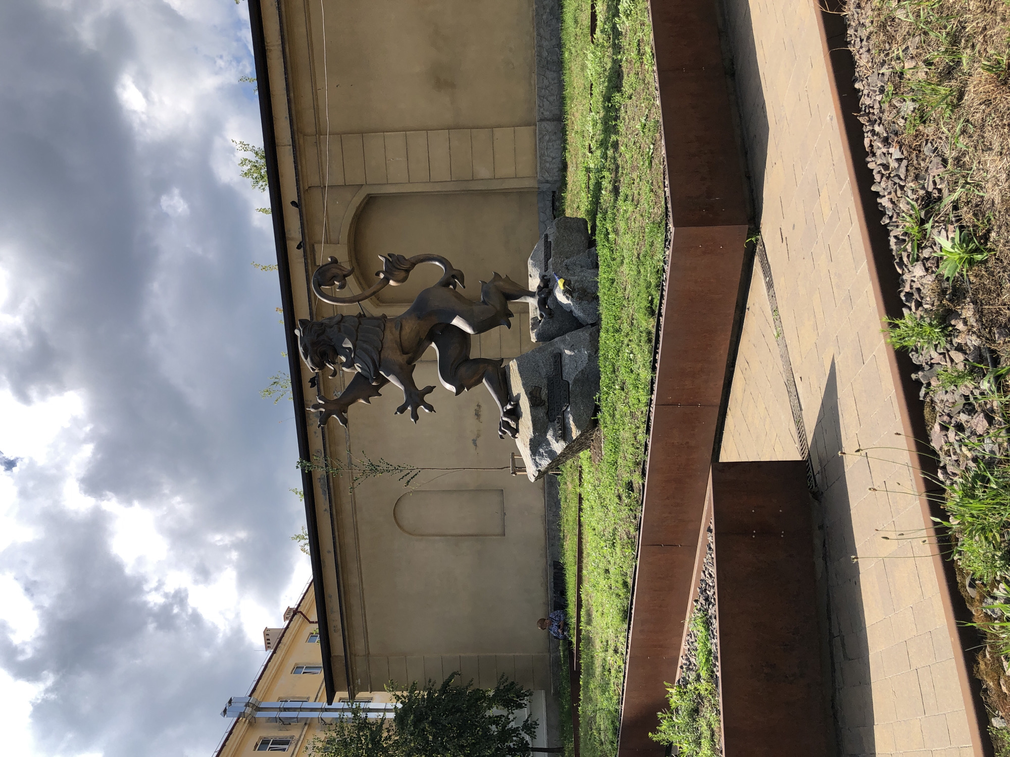 The image shows a bronze sculpture of a lion with intricate details, standing on rocks. The sculpture is positioned in an outdoor setting with a building in the background and a pathway made of beige pavers. Green grass and some gravel are visible around the sculpture and pathway, contained by rusted metal borders. The building in the background is light yellow with arched windows and some architectural details. There are a few people in the distance, one sitting on the grass. The sky is cloudy, suggesting it might be overcast or preparing for rain. The overall scene appears to be a public space, possibly a park or courtyard. The orientation of the image is rotated, with the sky appearing on the left side.