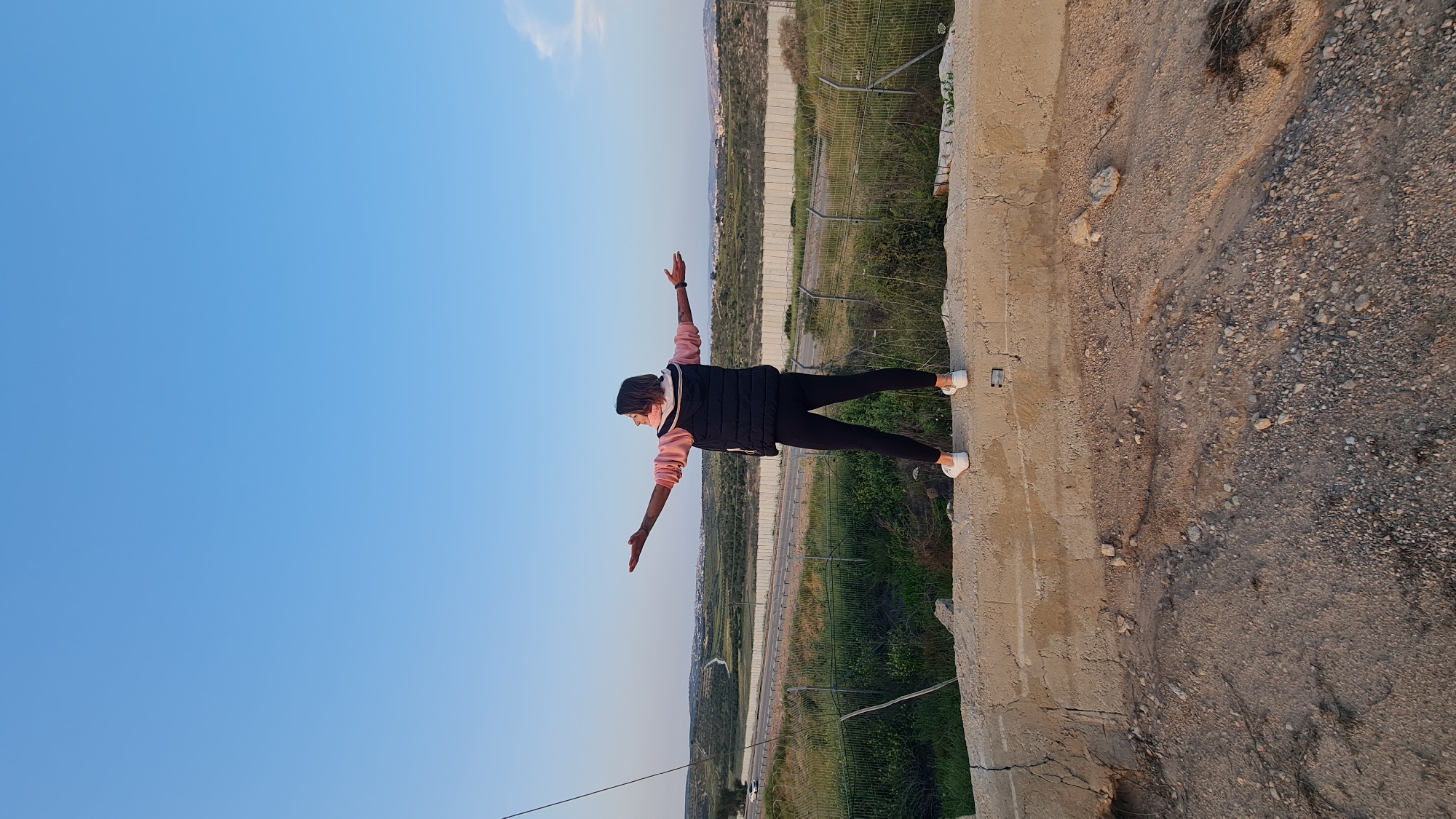 A person stands on a concrete structure in Lachish Regional Council, Israel, with arms outstretched against a background of the sky and landscape. The person wears a pink top, black vest, black pants, and white shoes. A fence runs parallel to a road in the distance. The ground is covered with dirt and gravel.