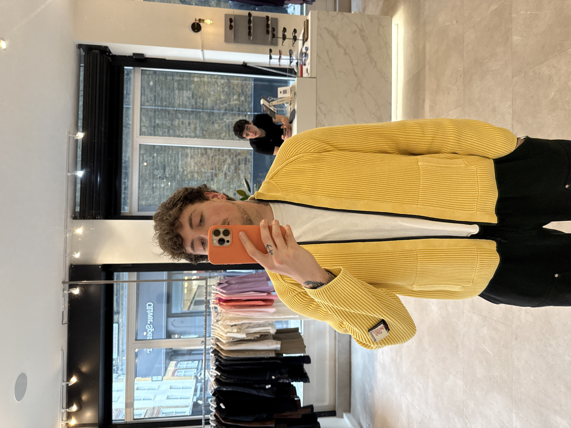 The image captures a mirror selfie taken by a young man inside a modern retail store, likely a fashion boutique. The man, with curly brown hair, is wearing a white t-shirt and a bright yellow, ribbed, zip-up cardigan or jacket, paired with black trousers. He holds an orange iPhone to take the photo, and tattoos are visible on his left hand and forearm, along with a ring on his right hand. A small, rectangular brand patch is present on the sleeve of his yellow jacket.
The store's interior is clean and well-lit, featuring light-colored walls and flooring. To the left, clothing racks display a selection of garments, including neatly folded items in pastel shades, white, and black, as well as clothes on hangers. In the background, a large window offers a view of an urban environment with brick buildings and a street. Reflected in the mirror, another young man in a black t-shirt can be seen leaning on a white counter, which also holds a display of sunglasses. The reversed text "Oliver Spencer" is visible through the window reflection, suggesting the name of the establishment or a neighboring business. The natural light indicates it is daytime. The overall setting is contemporary and suggests a stylish shopping environment in London.