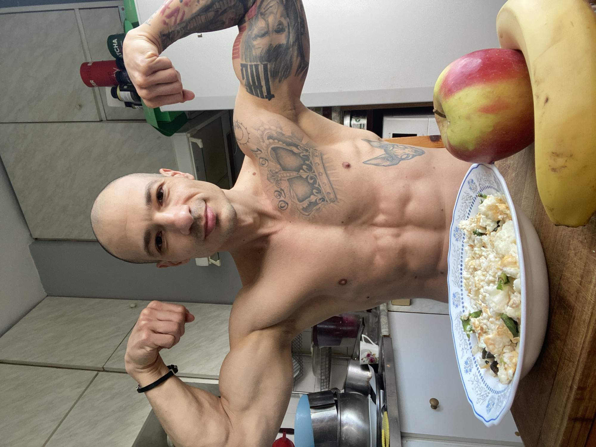 The image shows a bald, muscular adult male, shirtless, flexing both biceps with clenched fists, and looking directly at the camera with a slight smile. He exhibits prominent tattoos, including a large crown on his chest, an animal head on his right torso, and a detailed sleeve on his left arm featuring a face and geometric patterns. A black bracelet is visible on his right wrist.
In the foreground, on a wooden cutting board, rests a white plate with a blue decorative rim, holding a meal composed of a granular white substance (consistent with cottage cheese or scrambled eggs) mixed with green elements. Adjacent to the plate are a red and yellow apple and a yellow banana.
The background depicts a domestic kitchen environment. White upper cabinets are visible, along with a light gray countertop. To the left, a metal sink area contains stacked metal bowls and a white mug with a floral design. To the right, various containers, including a red one partially labeled "TER" and a green one partially labeled "ATCHA" (likely supplements), are on a shelf or cabinet surface, with a white appliance partially visible below. The specific city location of Pleven, Bulgaria, cannot be visually confirmed from the image content.