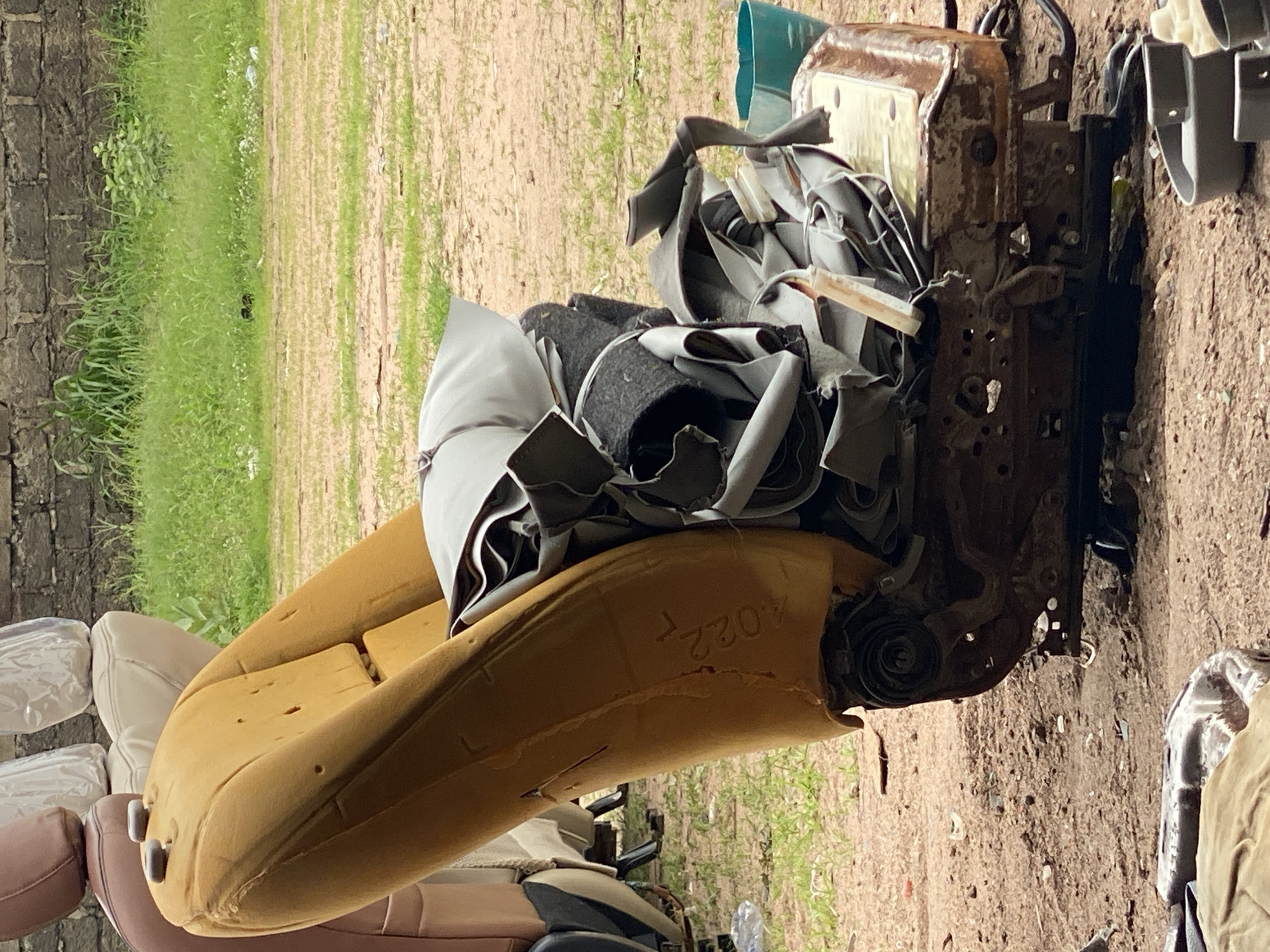 In Jalingo, Nigeria, a damaged car seat lies on the ground, its yellow foam padding exposed. The seat is partially disassembled, revealing its internal metal frame and springs. Various pieces of torn upholstery and padding are piled on top of the seat base. In the background, a brick wall and overgrown grass are visible. Several other car seats, some covered in plastic, are also present but out of focus. A teal plastic bucket is situated near the damaged seat. The ground is a mixture of dirt and sand with some scattered debris.
