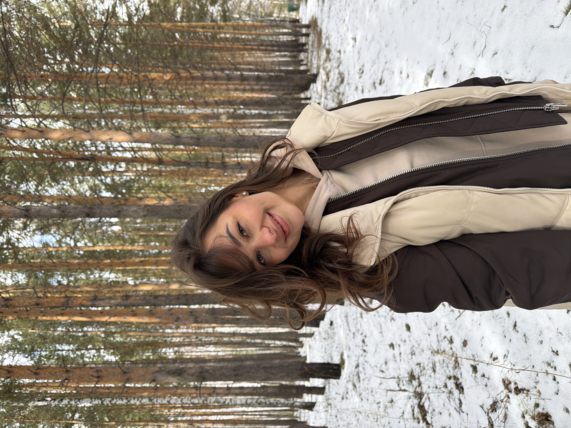 A young woman is photographed outdoors in a snowy forest. The scene captures a path lined with tall, slender trees, likely coniferous, which create a dense, vertical pattern extending into the distance. Snow covers the ground, interspersed with patches of dried grass. The woman, positioned in the foreground, wears a two-toned jacket in shades of brown and cream, and her dark hair is styled with loose curls. The photograph is taken at an angle, with the camera tilted to the side. Given the environment and the provided location of Krasnoyarsk, Russia, it is probable the photo was taken in a forested area surrounding or within the city.