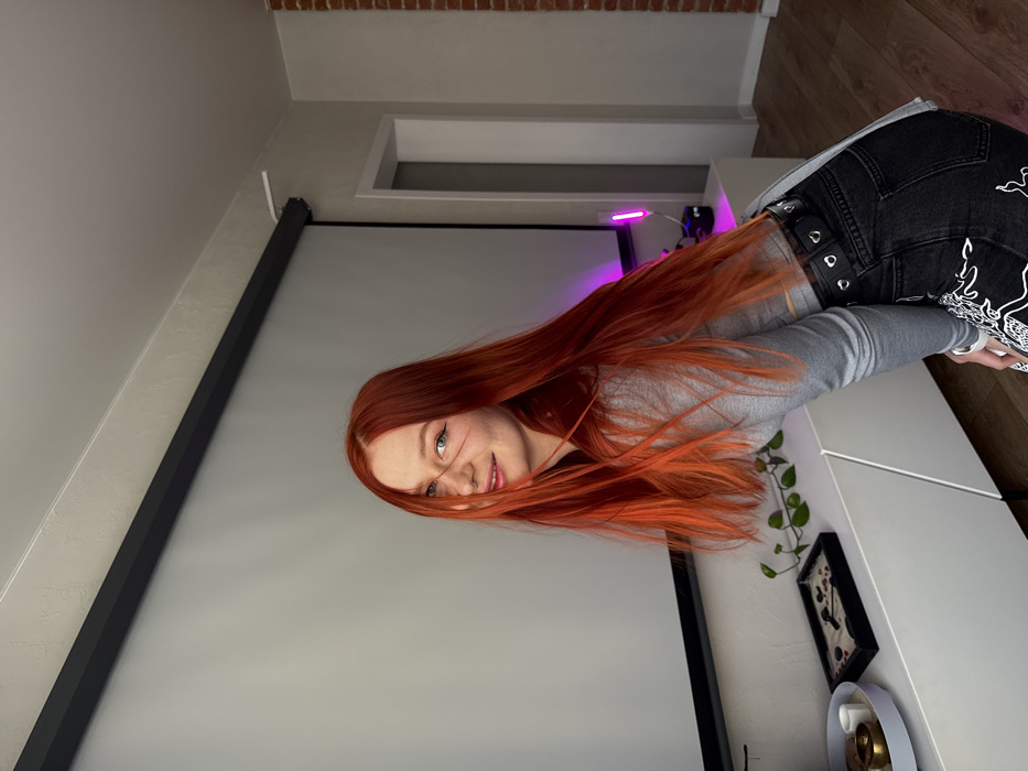 An individual with long, bright red hair and freckled skin, identified as female, poses in an indoor residential setting in what is described as Rostov-on-Don, Russia. She wears a light grey long-sleeved cropped top and black denim jeans adorned with white flame or abstract designs on the lower legs, secured by a black eyelet belt. A silver hoop nose piercing is visible on her left nostril. Her head is tilted slightly to the right, and her body is bent forward and to the left, as she looks directly at the camera with a slight smile.
Behind her, a large white retractable projector screen is partially extended on a wall. Below the screen, a long, low white media console or cabinet displays several items: a light grey tray with a small brass bowl and two white cylindrical objects on the far left, a black rectangular tray containing sand and dark elements (resembling a miniature zen garden) to its right, and a trailing green leafy plant. Further to the right on the cabinet, a vertical white light fixture emits a prominent pink-purple glow, with a dark electronic device positioned beneath it. A doorway is visible in the background to the right, leading into an adjacent room. The far right edge of the frame reveals a section of an interior wall constructed from exposed red brick. The floor consists of light-colored wood laminate. The ceiling is white.