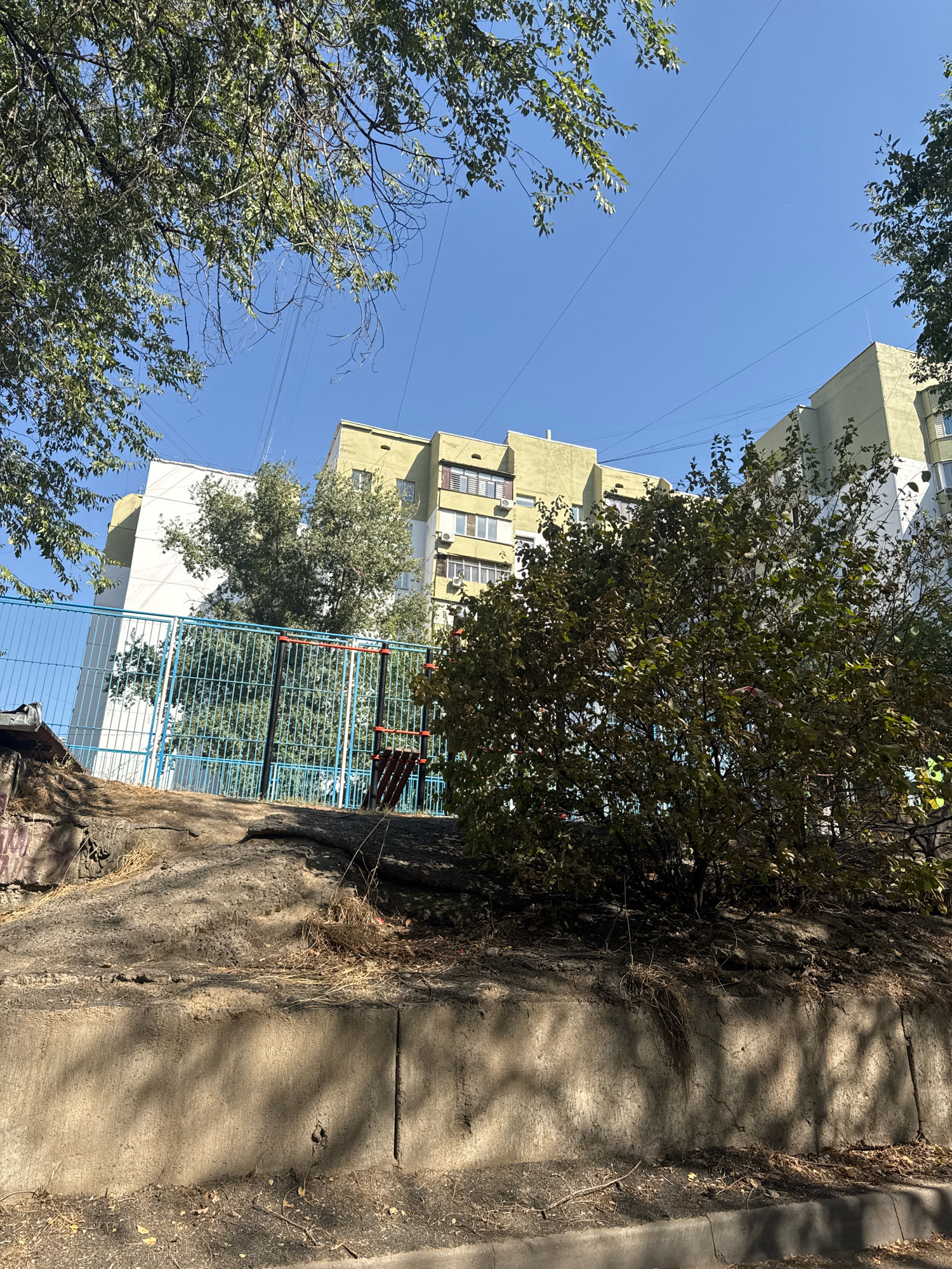 An outdoor, ground-level, wide shot shows a residential apartment building complex in Almaty, Kazakhstan, under a clear blue sky. The building, a multi-story structure with light yellow and white facades, has numerous windows and balconies. In the foreground, a rugged, dirt-covered embankment with patches of dry grass and some debris leads to a blue metal fence. Behind the fence, a section of a playground or outdoor gym with red and black bars is visible. Lush green trees with branches extending into the frame partially obscure the view of the building and the sky. The overall lighting suggests it is daytime, with shadows cast by the trees on the embankment and the lower portion of the building. The scene depicts a typical urban residential area.