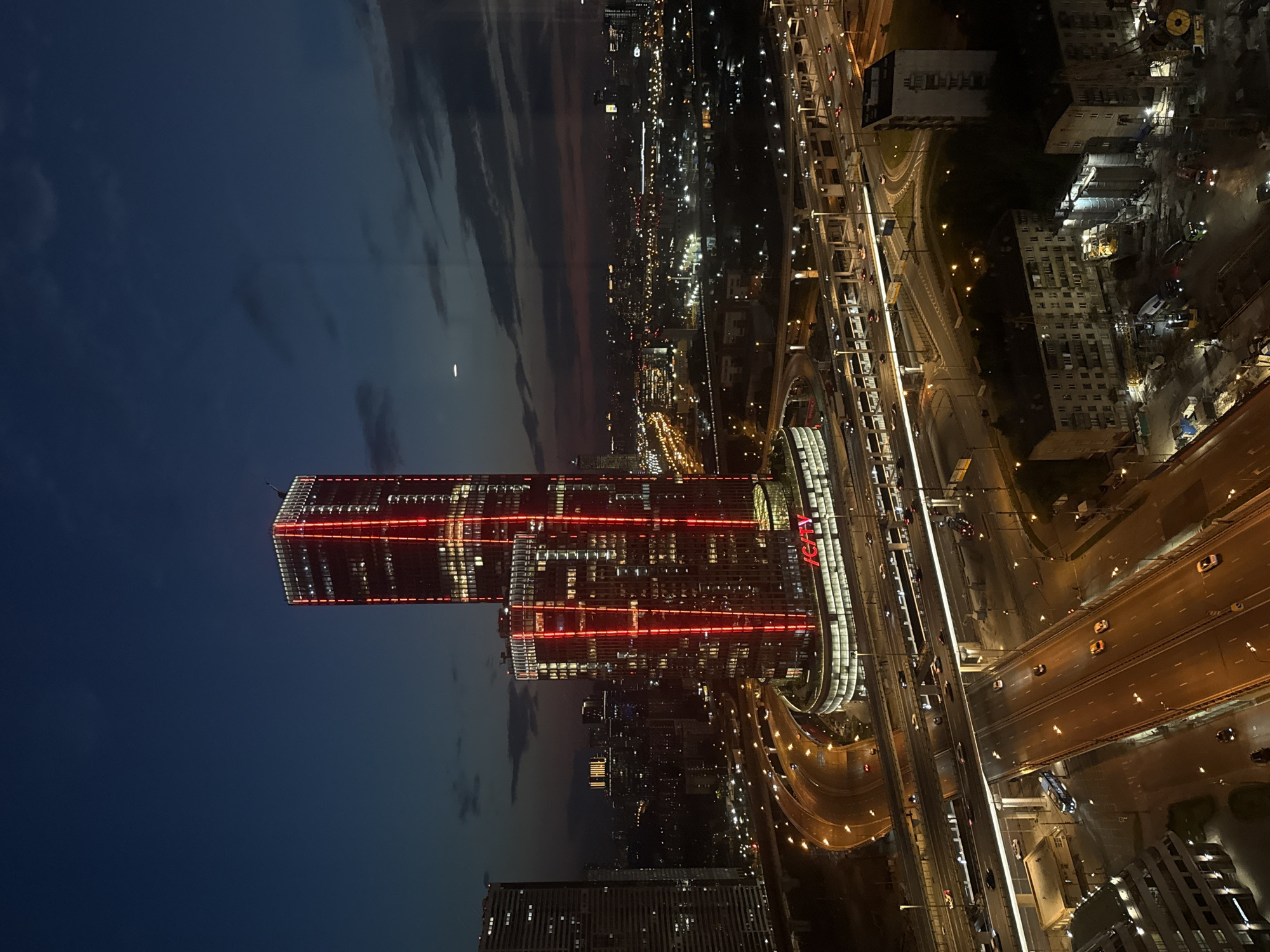 This is a nighttime aerial photograph of the city of Khimki, Russia, focusing on the prominent Icity Towers. The image captures a dense urban landscape characterized by a network of illuminated highways, multi-story buildings, and the striking architectural presence of the two skyscrapers. The Icity Towers are the central subject, their facades adorned with vertical red lighting that dramatically contrasts with the dark evening sky. The sky itself is a deep blue, with scattered clouds that are faintly illuminated by the city lights.
The foreground is dominated by a complex interchange of elevated roadways, with cars appearing as streaks of light due to the long exposure. The highways are brightly lit by streetlights, highlighting the infrastructure that connects different parts of the city. To the left and right of the Icity Towers, other tall buildings are visible, some with their windows lit, indicating occupancy. Further in the distance, the urban sprawl extends, marked by a multitude of smaller lights that form a glowing tapestry across the horizon.
In the lower right quadrant of the image, a construction site is visible. This area features cranes, construction equipment, and partially built structures, suggesting ongoing development within the city. The overall impression is one of a dynamic, modern city at night, with a focus on the bold design of the Icity Towers and the intricate transportation network surrounding them. There are no visible individuals in the photograph, and the actions depicted are limited to vehicular movement on the highways.