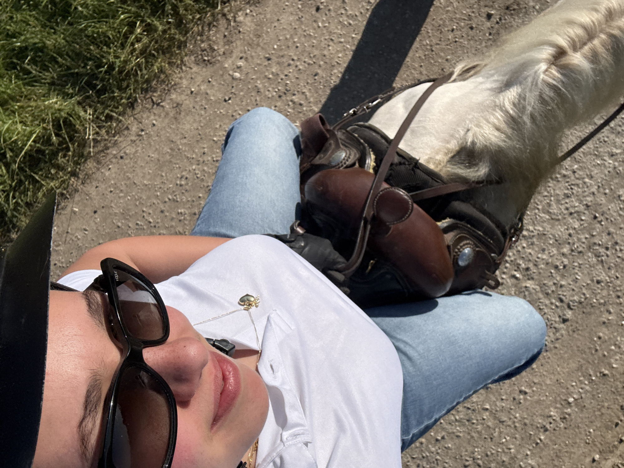 A woman wearing a white shirt, light blue jeans, and black sunglasses is sitting on a dirt path with a horse standing near her. The horse has a saddle on its back. Some grass is visible in the upper left corner of the image. The location is presumed to be the city of Spino d'Adda, Italy.