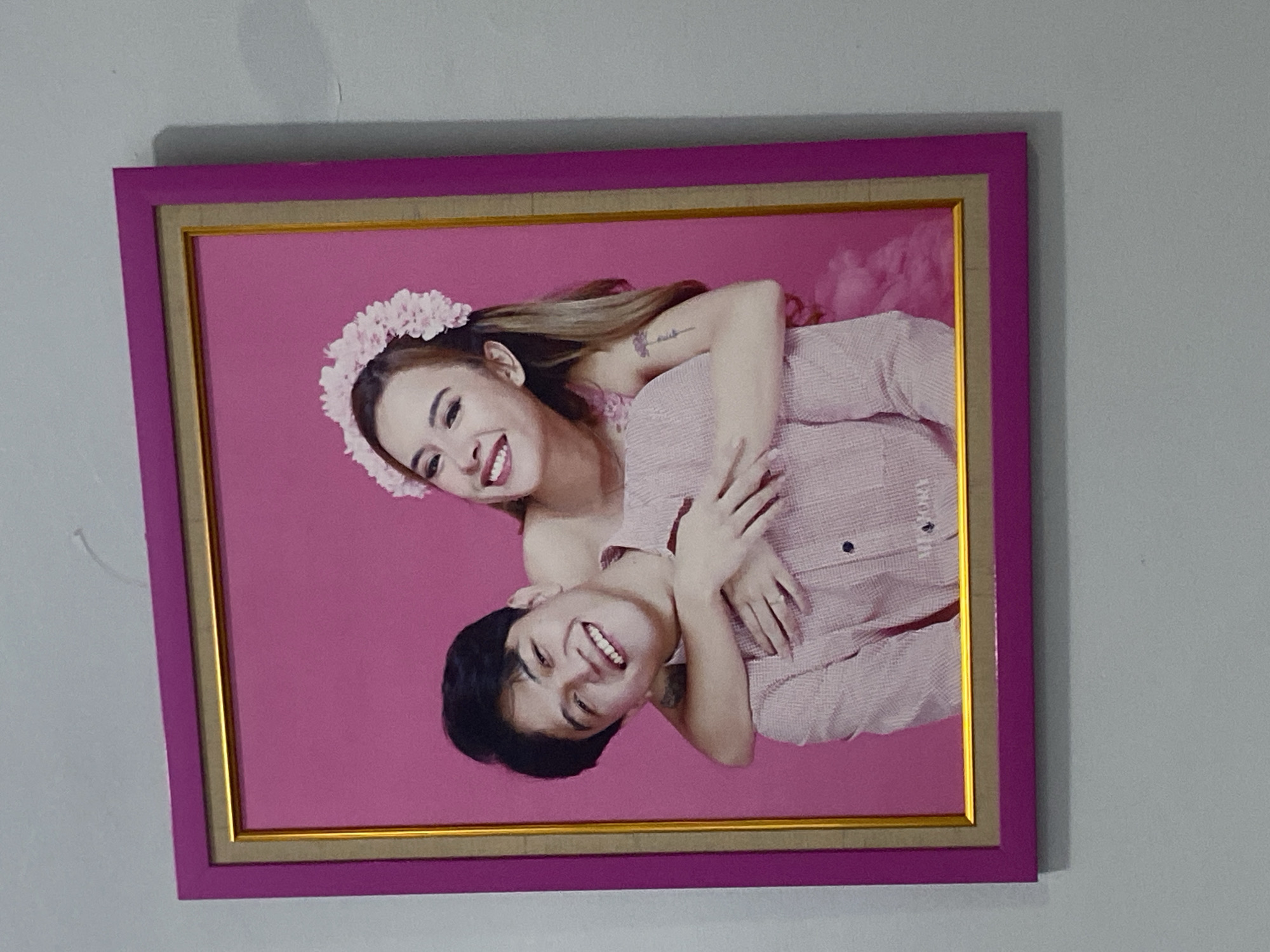 A framed portrait of a smiling young couple is displayed on a light gray wall. The photograph features a young man with short, dark hair, wearing a light pink collared shirt. He is looking at the camera and smiling. A young woman with long, brown hair adorned with a pink floral headband is embracing him from behind. She is also smiling broadly and has her arms wrapped around his neck and chest. The woman is wearing a pink dress. The background of the photograph is a solid, vibrant pink. The frame is a bright pink with a gold inner border. The image is hung on a wall in a location that appears to be a residential setting, potentially in Dukuh, Indonesia, given the context. The wall is plain and unadorned, except for the framed photograph.