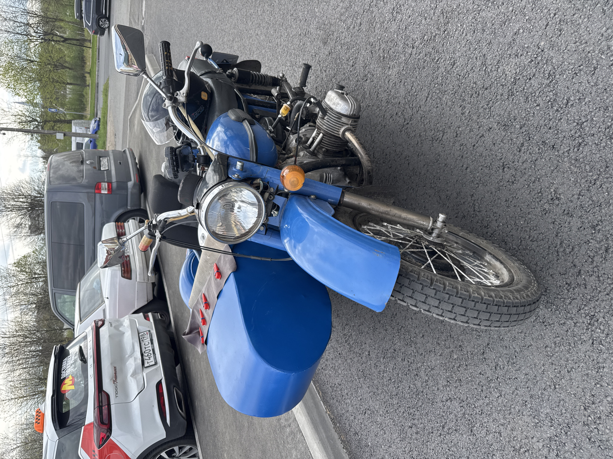 A bright blue motorcycle with a sidecar is parked on asphalt in an urban setting, potentially Saint Petersburg, Russia. The motorcycle has a visible headlight, an orange turn signal, and a sidecar with a gray cloth bearing small red objects inside. The background shows a taxi and other parked vehicles, along with some trees in the distance. The day appears to be overcast with gray skies.