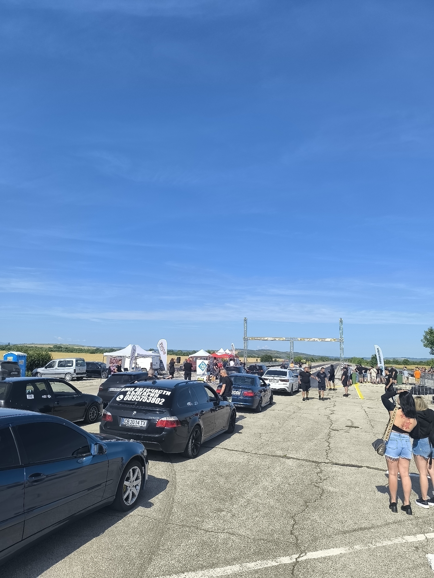 The image depicts an outdoor event, likely a car show or race, on a sunny day. The location is identified as the Akatsievo, Bulgaria, drag strip. In the foreground, several cars are parked on a paved surface. A dark blue sedan is prominently featured on the left, with its wheels and side visible. Behind it, a black BMW wagon is parked, displaying a Bulgarian license plate "CB 2014 TX" and text on its rear window. Further back, other vehicles and tents are visible. A group of people are gathered near a start/finish line structure, with some walking and others standing. In the background, a field of wheat or similar crop stretches towards a treeline and rolling hills under a clear blue sky with scattered clouds. The scene appears to be a public event with attendees observing or participating in activities related to cars.