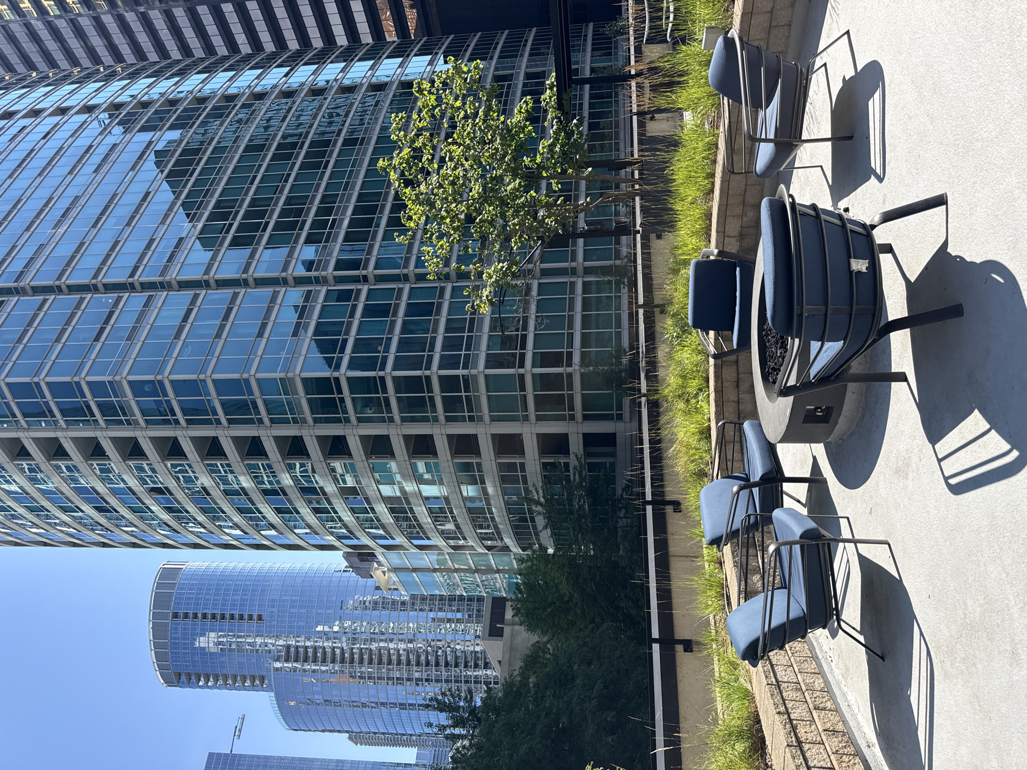The image depicts a modern rooftop patio in Chicago, United States of America, featuring a fire pit surrounded by four blue upholstered chairs with black metal frames. The patio is paved with concrete and bordered by a low stone wall topped with ornamental grasses. Behind the patio, a large, contemporary high-rise building with a glass facade dominates the scene, its windows reflecting the bright blue sky. To the left, other skyscrapers with similar architectural styles are visible, adding to the urban landscape. A lone, leafy tree stands in the middle ground, casting a shadow on the patio. The overall impression is one of a serene urban oasis high above the bustling city.