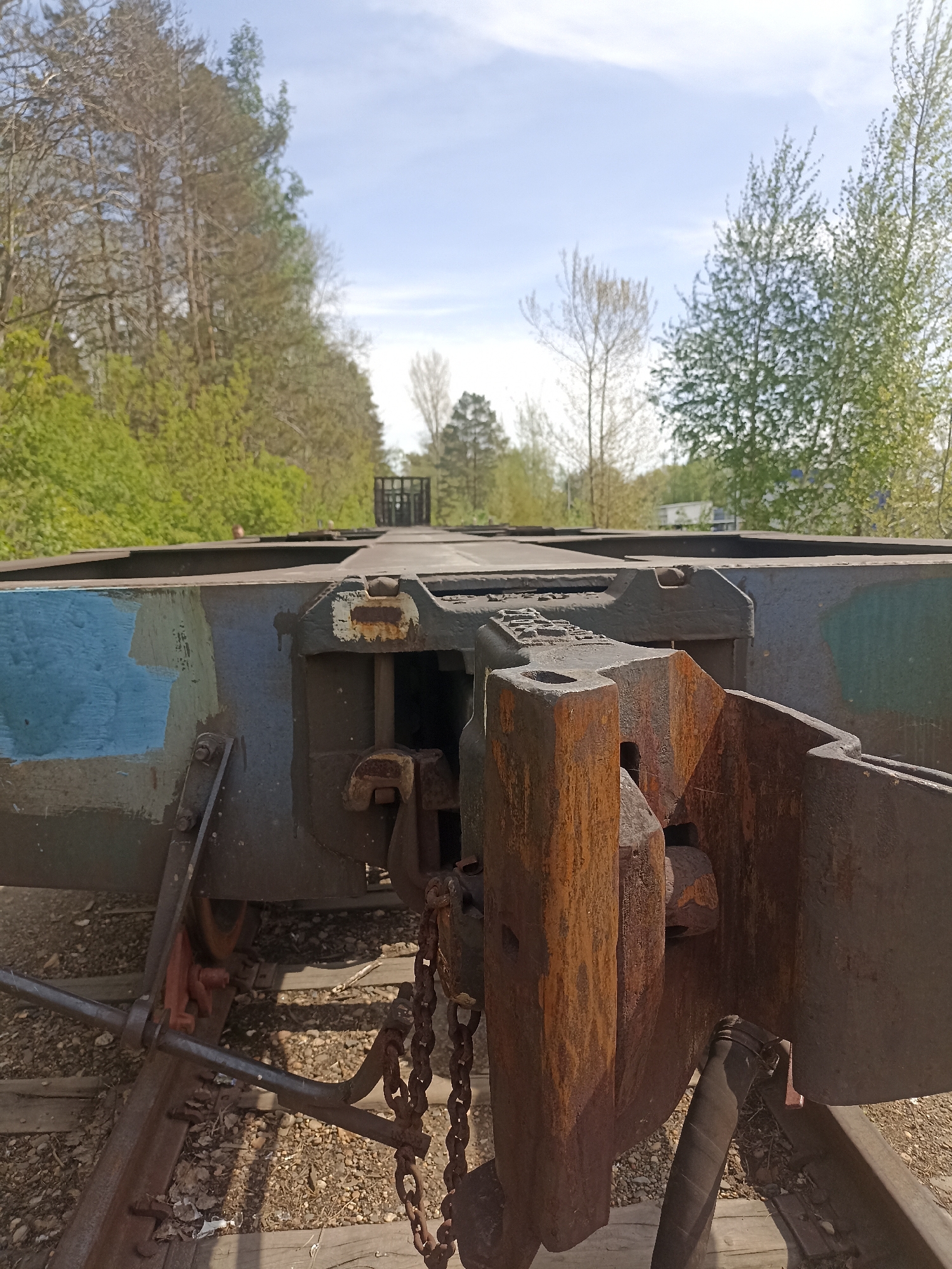 The image shows the coupling mechanism of a railcar, heavily rusted, in an outdoor setting. The railcar is on a track, with vegetation visible on either side. The sky is visible in the background. The location is not determinable from the image.