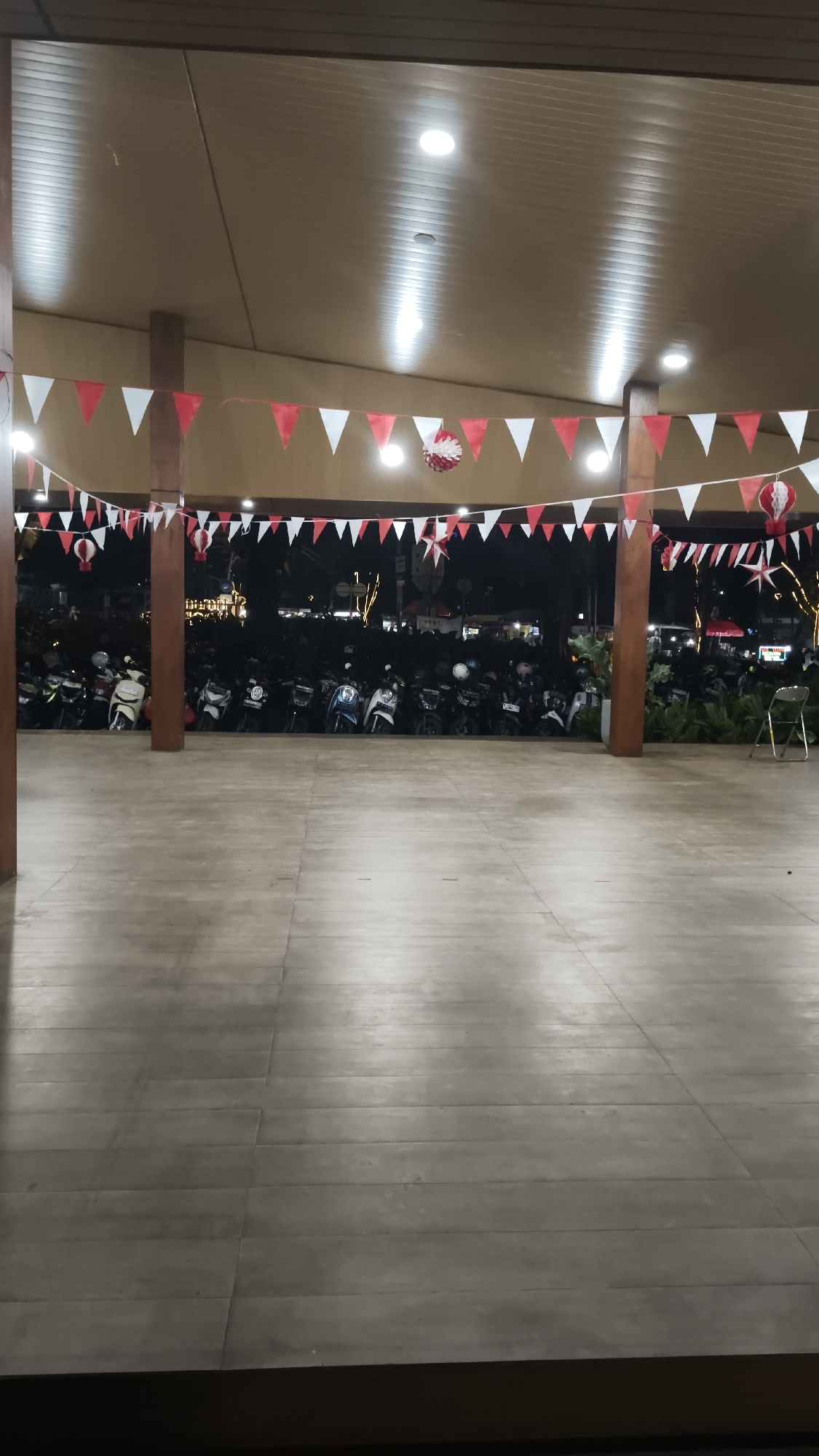 A long shot captures an outdoor area at night, likely a parking lot or public space under a covered overhang. Red and white pennant banners are strung across the ceiling of the overhang, with decorative red and white spherical ornaments hanging among them. Below the overhang, a multitude of motorcycles are parked in rows, filling the space from the foreground to the background. The ground is paved with large, light-colored rectangular tiles. Beyond the parked motorcycles, faint streetlights and building lights are visible, indicating the presence of a surrounding urban environment. A single, empty silver folding chair is placed on the right side of the image, near the edge of the covered area. The scene appears to be in a city, given the lights and parked vehicles.