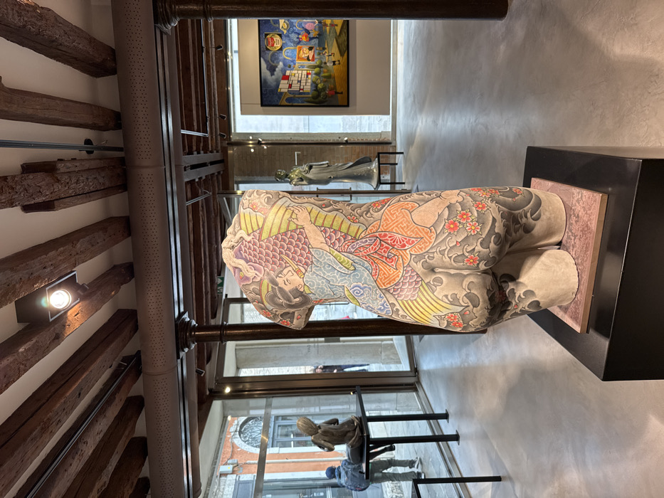 The image displays the interior of an art gallery or exhibition space located in Venice, Italy. In the immediate foreground, a light-toned, full-torso sculpture, appearing to be crafted from stone or concrete, is positioned on a black rectangular pedestal topped with a reddish square base. The sculpture is extensively adorned with a colorful, Japanese-style tattoo design covering its entire surface, depicting a figure in traditional attire embracing a large, scaled fish or dragon, surrounded by waves and cherry blossoms on the lower back and buttocks.
Through large glass panels on the left, an exterior Venetian street scene is visible. A person wearing a blue jacket and a backpack is partially seated near a dark, abstract sculpture of a seated figure, which rests on a black rectangular table. Another individual in dark clothing is indistinctly visible further along the street. The external architecture shows multi-story buildings with light-colored plaster and reddish accents, consistent with Venetian structures.
Within the gallery, towards the mid-right, a standing figure sculpture, light in color and possibly ceramic, is displayed on a pedestal. In the far right background, a large, brightly colored framed painting hangs on a light grey wall, depicting a stylized landscape with architectural elements, small figures, and prominent Pop Art imagery including a "Campbell's Soup" can.
The gallery ceiling features exposed dark wooden beams with embedded modern track lighting fixtures. A long, light-colored, perforated ventilation duct runs horizontally below the beams. Several dark, fluted metal columns support the overhead structure. The floor is a smooth, polished light grey concrete.