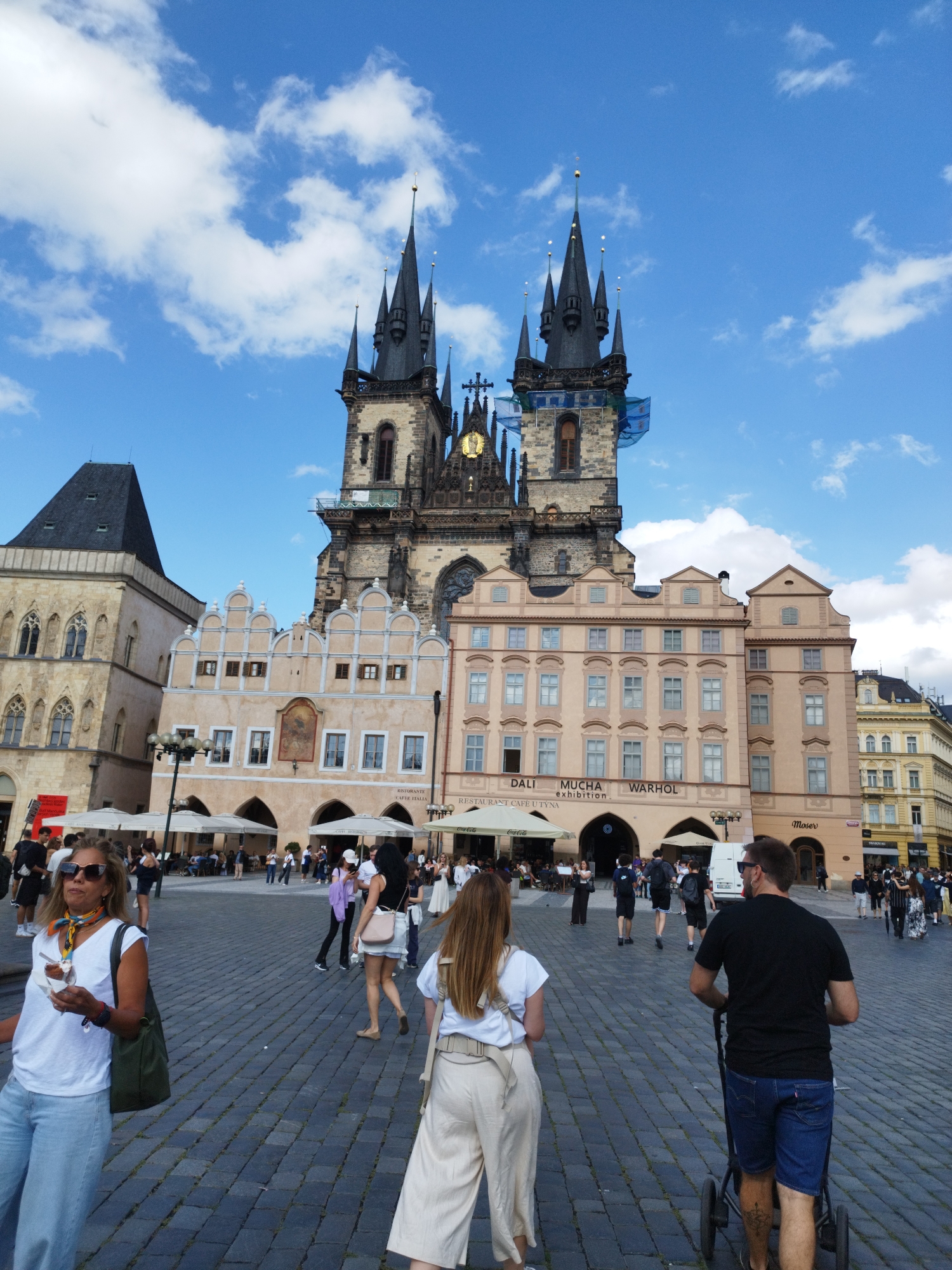This is a medium shot, eye-level, full-color outdoor photograph taken on a cobblestone square in front of a Gothic church with two prominent spires. The sky is blue with scattered white clouds. In the foreground, several people are walking. A woman in the left foreground wears a white t-shirt, light blue wide-leg pants, and sunglasses, and carries a dark green bag over her shoulder. She holds a piece of food in her right hand. To her right and slightly behind her, a woman in a white t-shirt and tan cropped pants walks away from the camera, her back to the viewer. Further back and to the right, a man in a black t-shirt and blue shorts pushes a stroller, his back to the camera. He wears sunglasses and has a tattoo on his right calf. In the middle ground, a crowd of people are walking and standing, some in front of the church's facade. The church has a large clock face visible between its spires. The buildings surrounding the square are a mix of architectural styles, with arched doorways and windows. Signs for "DALI MUCHA WARHOL exhibition" and "RISTORANTE CAFFE ITALIA" are visible on the buildings. The overall scene depicts a busy public square on a bright, partly cloudy day.