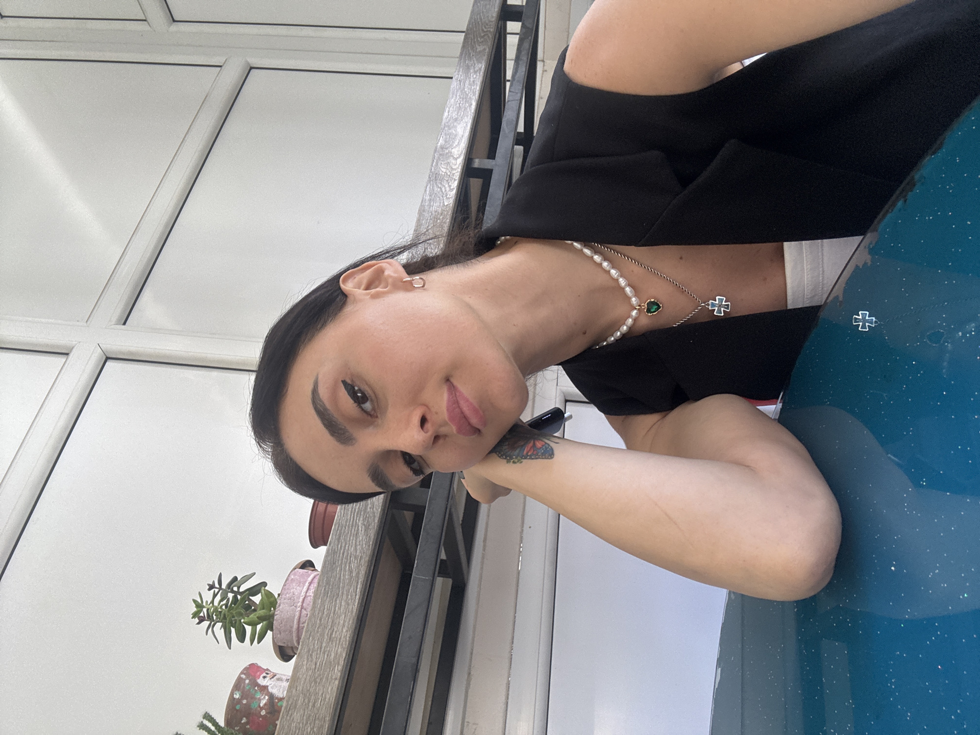 In this close-up, eye-level, indoor photograph, a young woman with dark hair and brown eyes leans her head on her left arm, which is resting on a dark teal surface. She is wearing a black vest over a white top and a pearl necklace with a green heart pendant, as well as a silver necklace with a cross pendant. Her right arm is visible, bent at the elbow. Her left arm has a butterfly tattoo. The background is out of focus but appears to show a window with white panes and shelves with small potted plants. The scene is brightly lit, possibly by natural light. The presence of plants suggests an indoor environment with some natural light access, potentially a cafe or a home. The woman's gaze is directed towards the camera. The location of Tbilisi, Georgia is not discernible from the visual information present in the image.