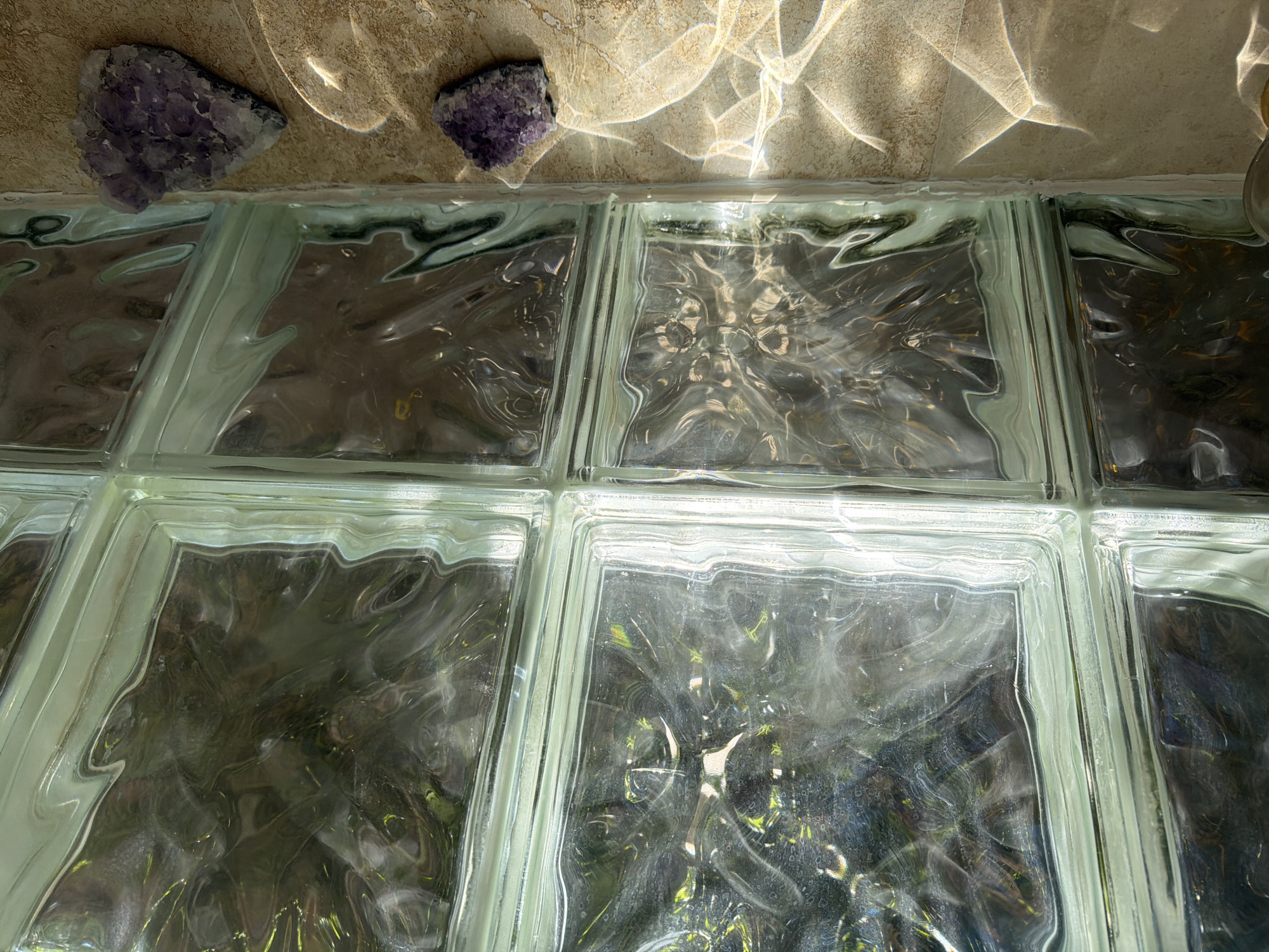 In Riviera Beach, Florida, USA, the image captures a close-up view of light passing through a wall of clear glass blocks. The glass blocks are arranged in a grid pattern, and the light refracts and distorts the view of the foliage on the other side, creating a dappled and wavy effect. The sunlight also casts intricate, shimmering patterns on the beige-tiled surface below the glass block wall. Two clusters of amethyst crystals are placed on the tiled surface, one to the left and one to the right, catching the light and adding a touch of color and texture to the scene.