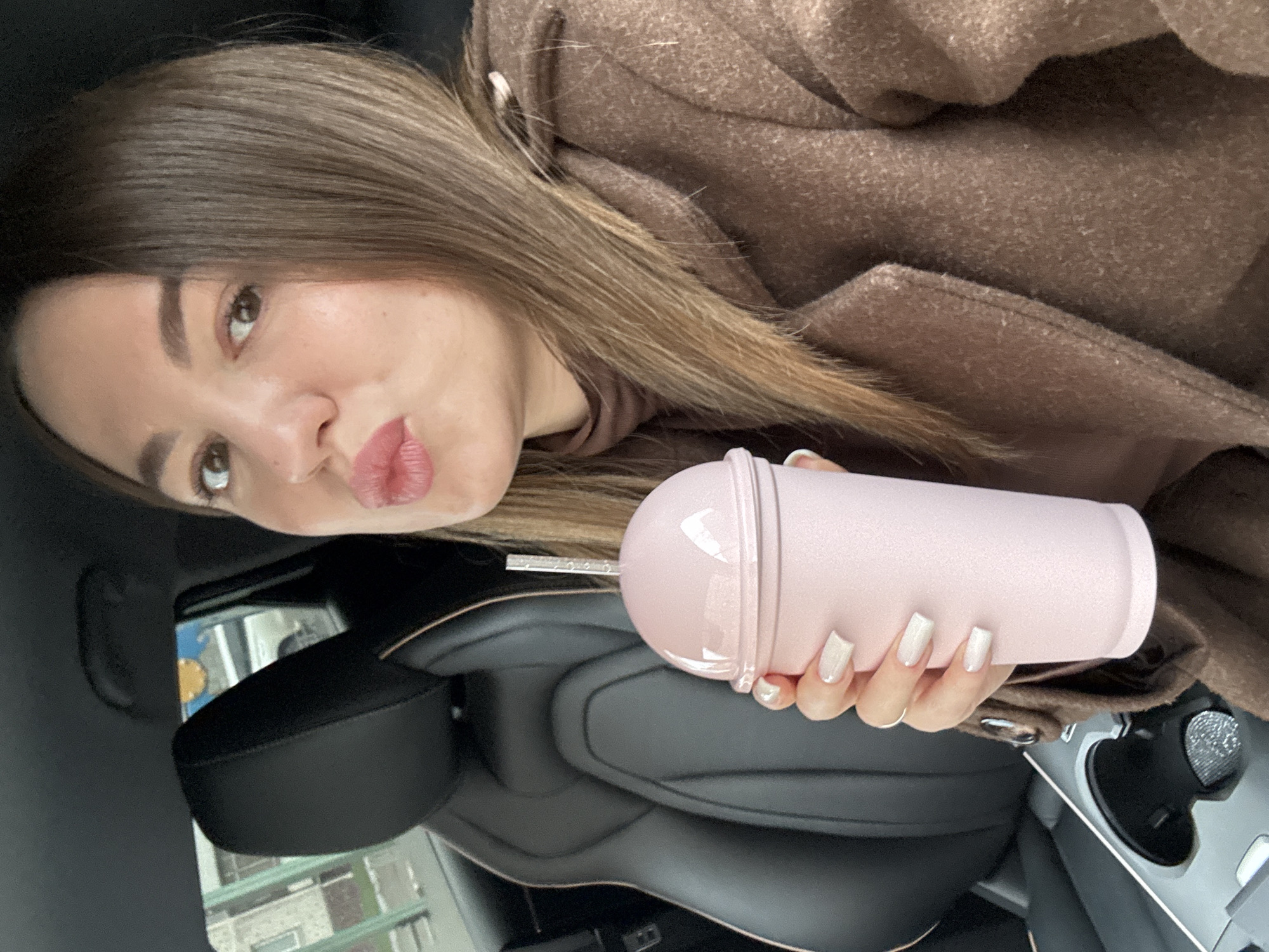 A woman is in the driver's seat of a car, holding a pink cup with a straw. She is wearing a brown coat and has brown hair. The car is likely parked in Likino-Dulyovo, Russia, based on street sign visible in the reflection on the windshield.