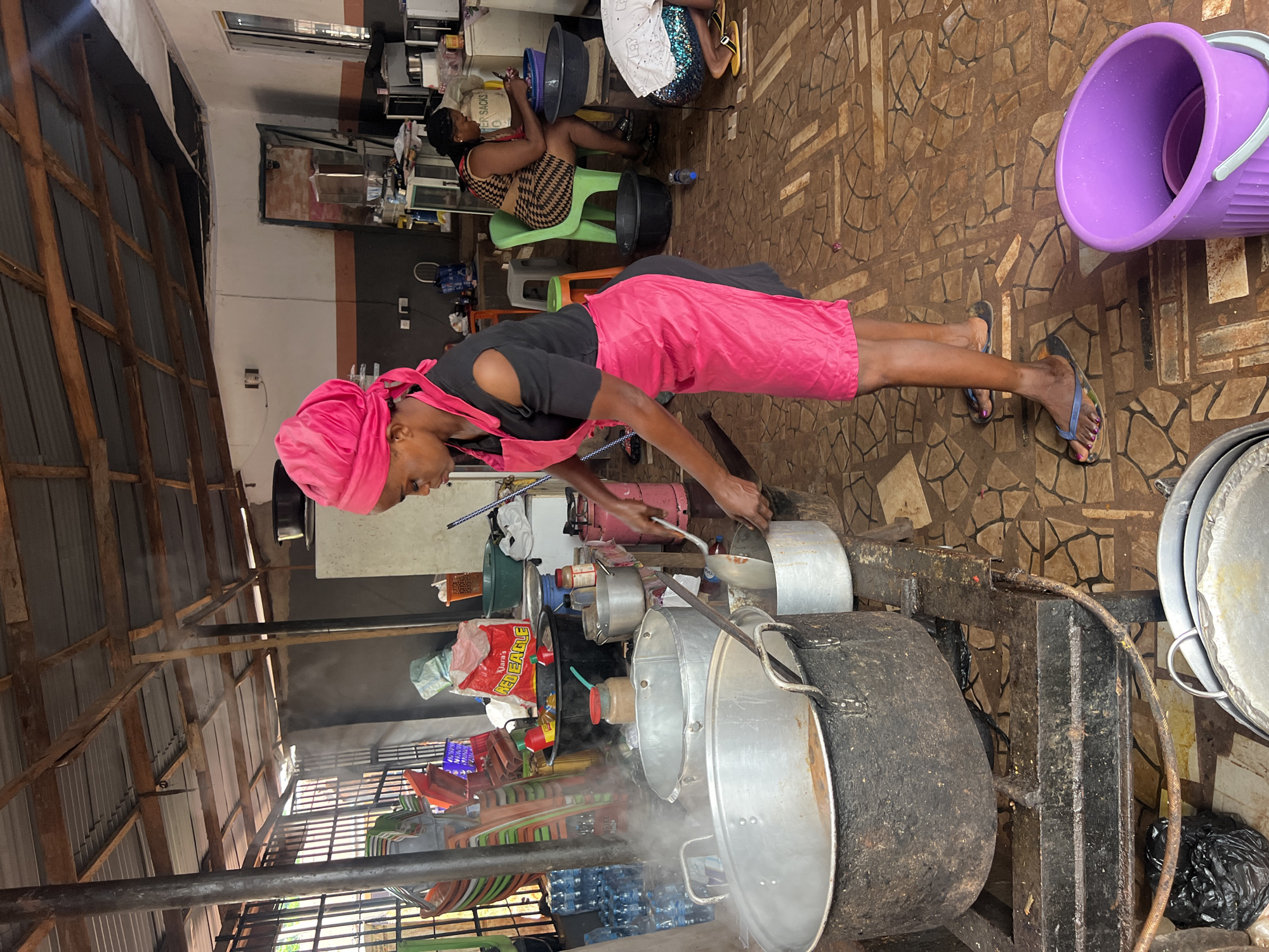In Nkpor, Nigeria, a woman wearing a pink headscarf and a pink apron over a black shirt is cooking in an open-air kitchen. She is stirring a pot with a ladle. There are several large metal pots on a metal stove, with steam rising from them. In the background, another woman is seated at a table, looking at her phone. The floor is tiled with a mosaic pattern. There are stacks of plastic chairs, water bottles, and various kitchen supplies visible. The ceiling is made of corrugated metal and exposed wooden beams.