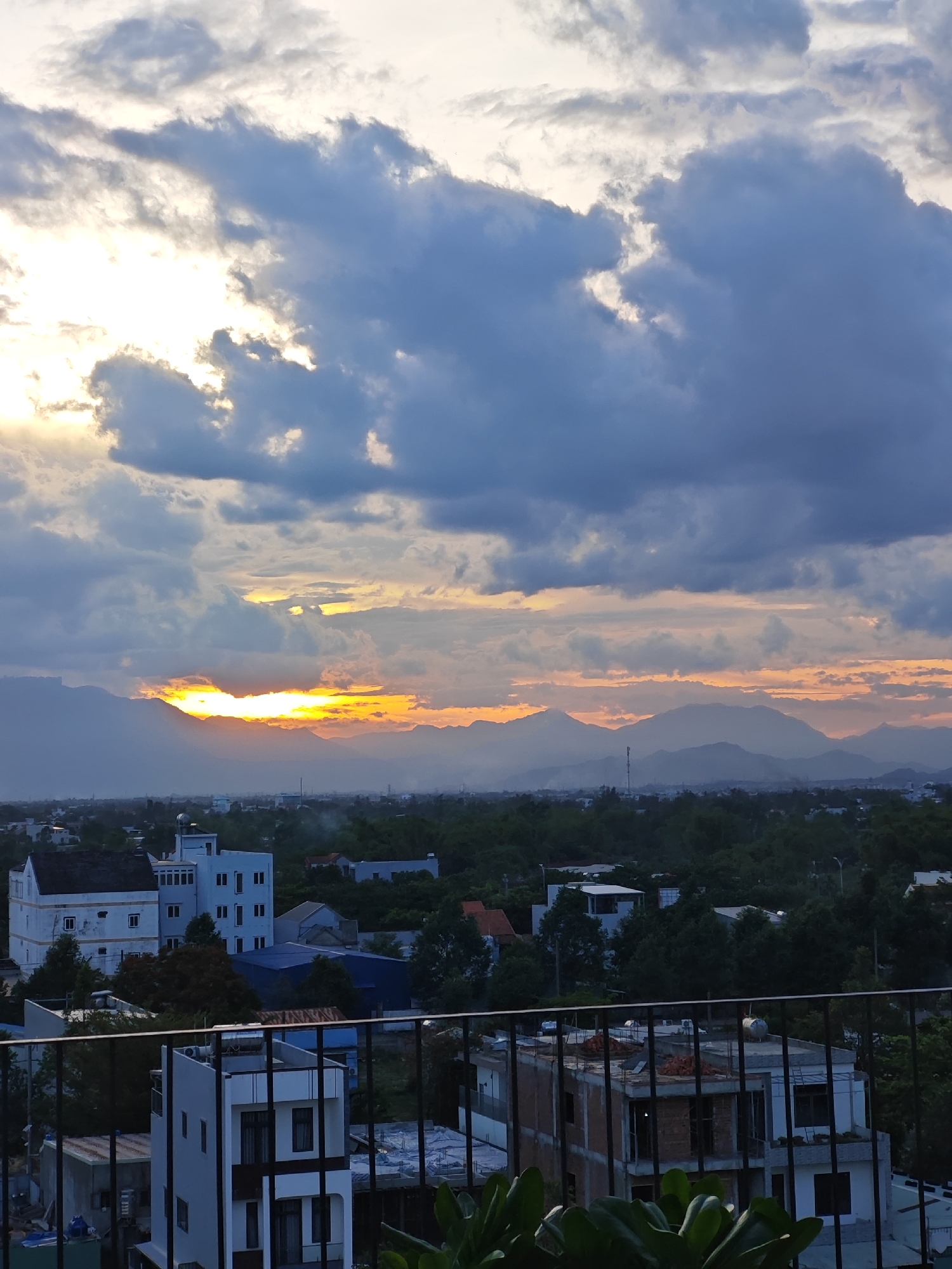 The image captures a sunset over a city in Đà Nẵng, Vietnam, as viewed from a rooftop. The sky is dramatic with dark, heavy clouds interspersed with bright, orange and yellow hues of the setting sun peeking through. In the distance, a range of hazy mountains forms a silhouette against the colorful sky. The mid-ground shows a dense urban landscape with numerous buildings, rooftops, and lush green trees. Closer to the foreground, there is a black metal railing, suggesting the vantage point is from a balcony or rooftop terrace. The buildings visible are a mix of modern architecture, including multi-story structures with white and grey facades, and some under construction. The overall atmosphere is one of a serene, yet vibrant urban environment at dusk.