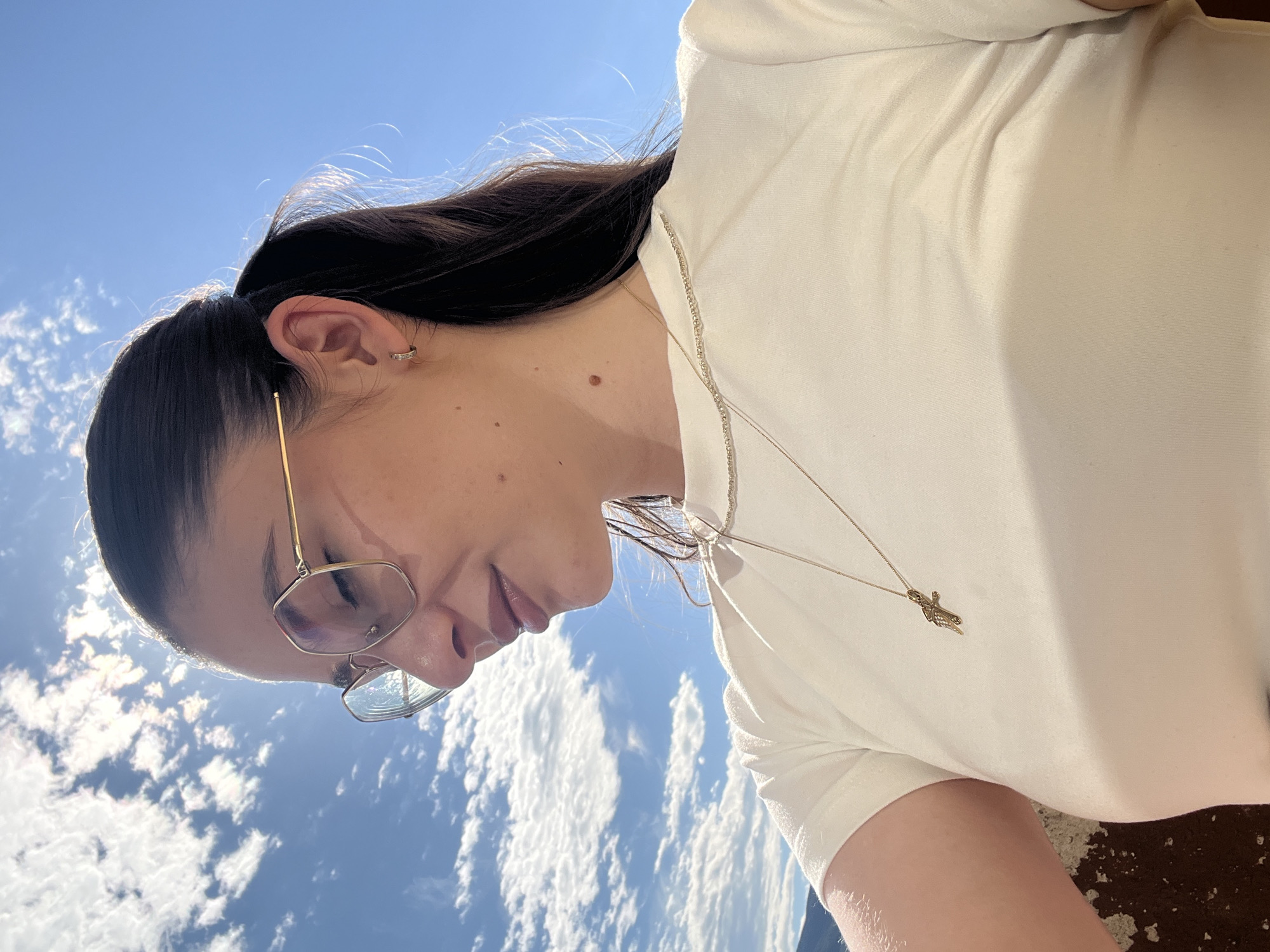 A close-up, outdoor, medium shot shows a young woman wearing a white t-shirt and gold-rimmed glasses. The woman has dark hair pulled back into a ponytail, and is wearing a small gold hoop earring in her left ear. A thin gold chain with a small crucifix pendant is visible on her chest. Her face is angled away from the camera, with her eyes closed. The background is a bright blue sky with scattered white clouds, and sunlight filtering through the clouds. A hint of a mountainous landscape is visible in the lower left corner.