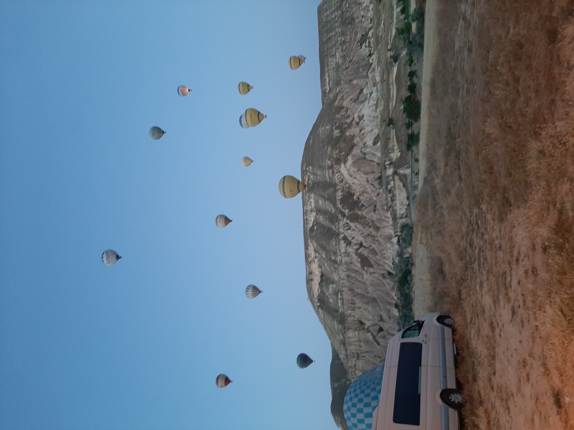 In Avanos, Turkey, a fleet of hot air balloons fills the pale blue pre-dawn sky over a rugged, mountainous landscape. The balloons, in various colors and patterns, are ascending or already airborne. In the foreground, a light-colored minivan is parked on a dirt and dry grass field. The terrain consists of dry vegetation and sandy soil, with a distinctive layered rock formation forming the backdrop. A blue and white checkered dome, possibly part of a structure, is visible on the left side of the image. The scene suggests an early morning hot air balloon launch, a common tourist activity in Cappadocia.