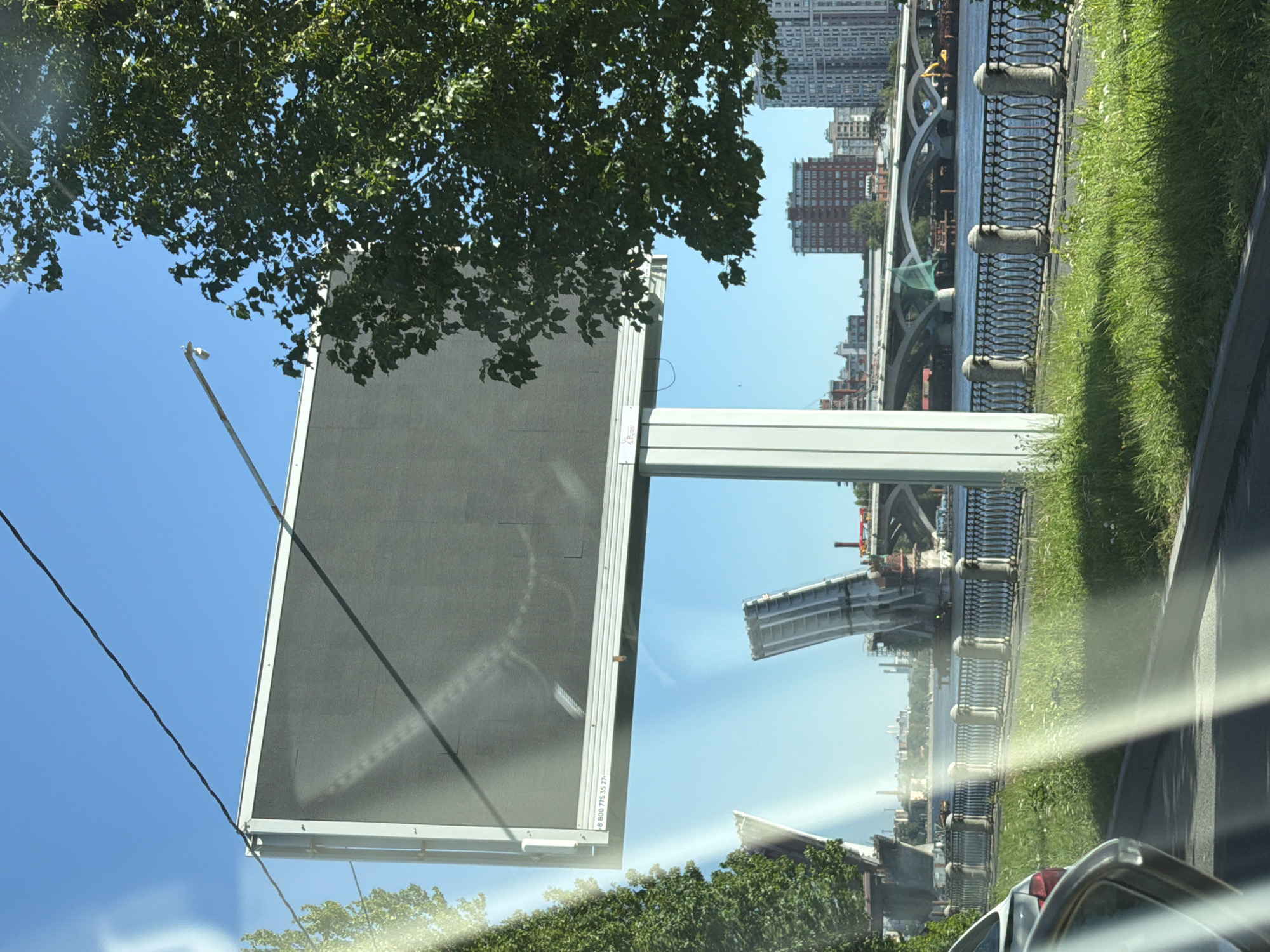 A wide, digital billboard is mounted on a tall, white pole in Saint Petersburg, Russia. The billboard is dark and appears to be off. A tree with green leaves partially obscures the upper right corner of the billboard. In the background, a large, modern bridge spans a body of water. To the left of the billboard, a bascule bridge is in the process of being raised. The scene is bathed in sunlight, casting shadows and reflections on the road and the billboard. A fence with decorative ironwork runs along the water's edge. Several tall buildings are visible in the distance. A car is partially visible in the foreground, suggesting the photo was taken from inside a vehicle.