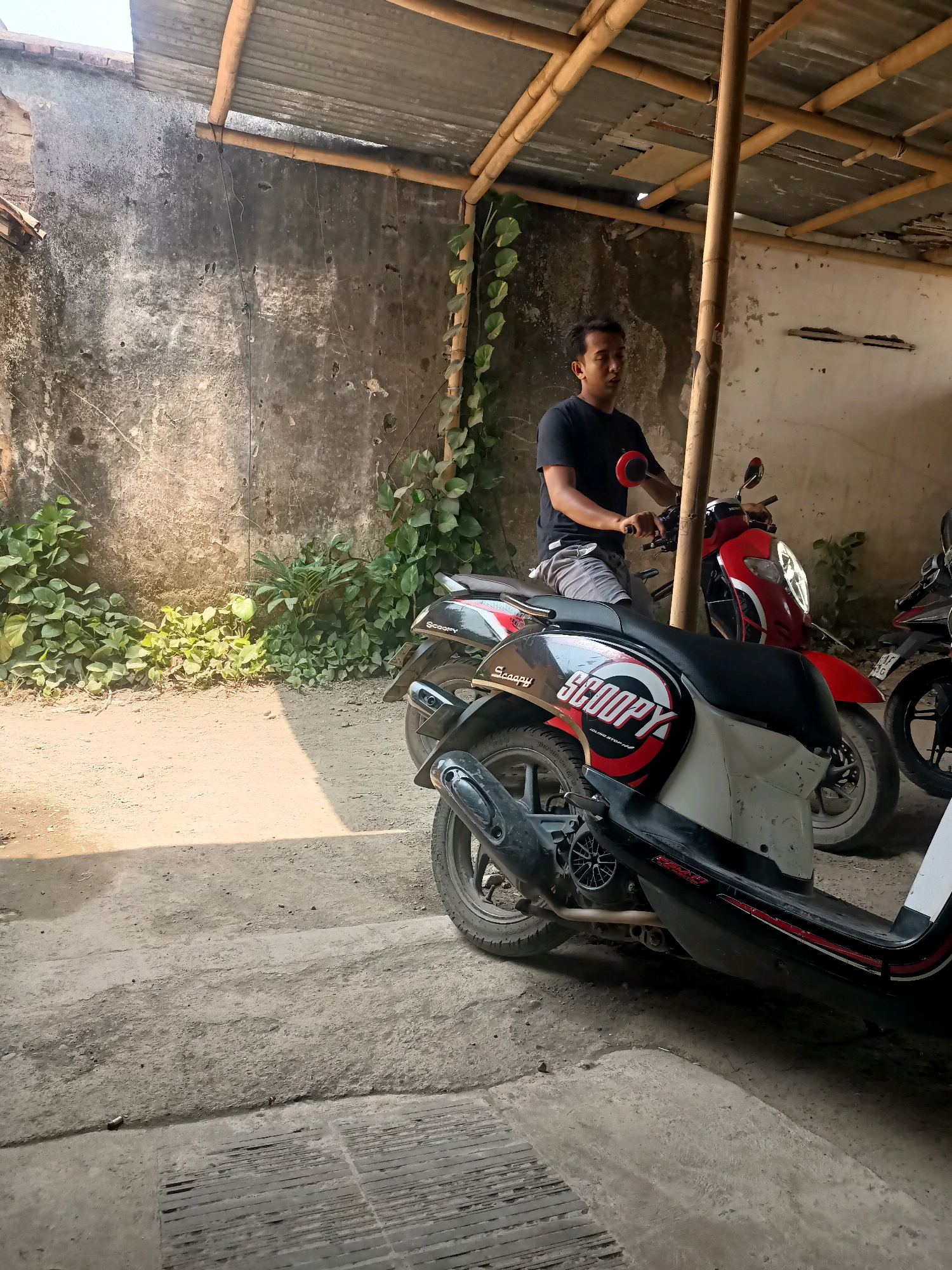 A person is sitting on a black and white scooter under a bamboo-supported corrugated metal roof in Tembok Luwung, Indonesia. The person is wearing a black t-shirt and grey shorts and is looking to the left. A red scooter is parked to the right of the black and white scooter, and another partially visible scooter is to the far right. The background consists of a rough concrete wall with vines growing on it, and the ground is a mixture of dirt and concrete. Sunlight is casting shadows on the ground.