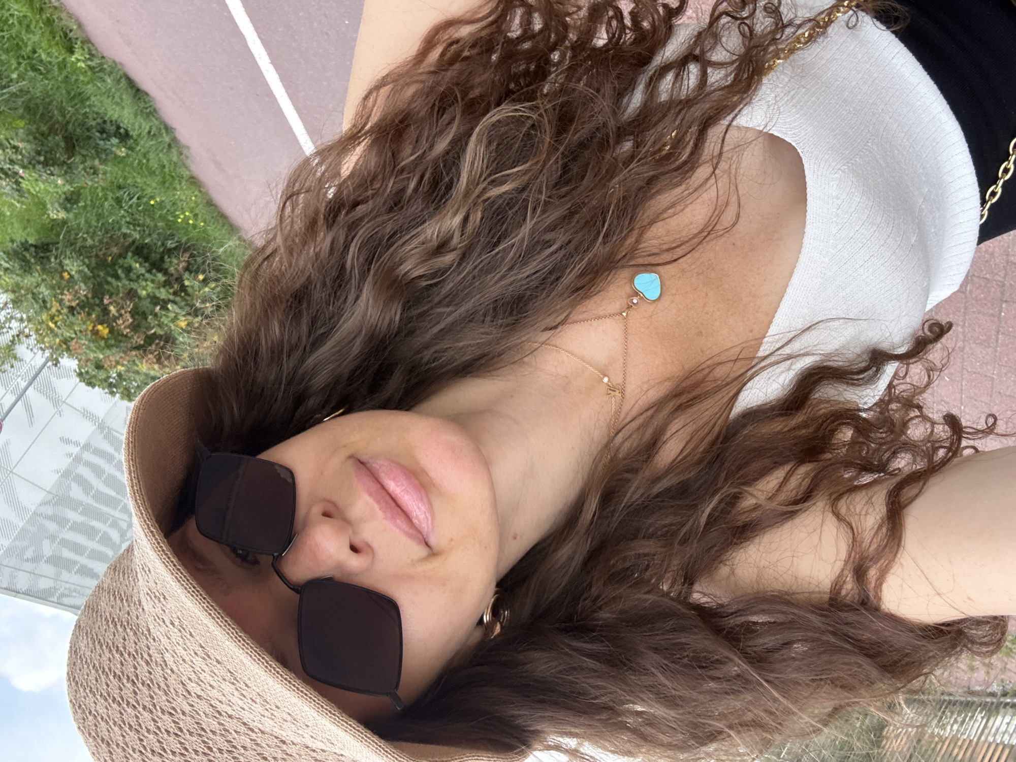 A woman wearing a straw hat and sunglasses takes a selfie outdoors. She has long, curly brown hair and is wearing a white top and a gold necklace with a turquoise heart pendant. The background shows greenery and a pink pathway, suggesting a park or outdoor recreational area. The image does not contain any specific indicators to place it in Moscow, Russia.