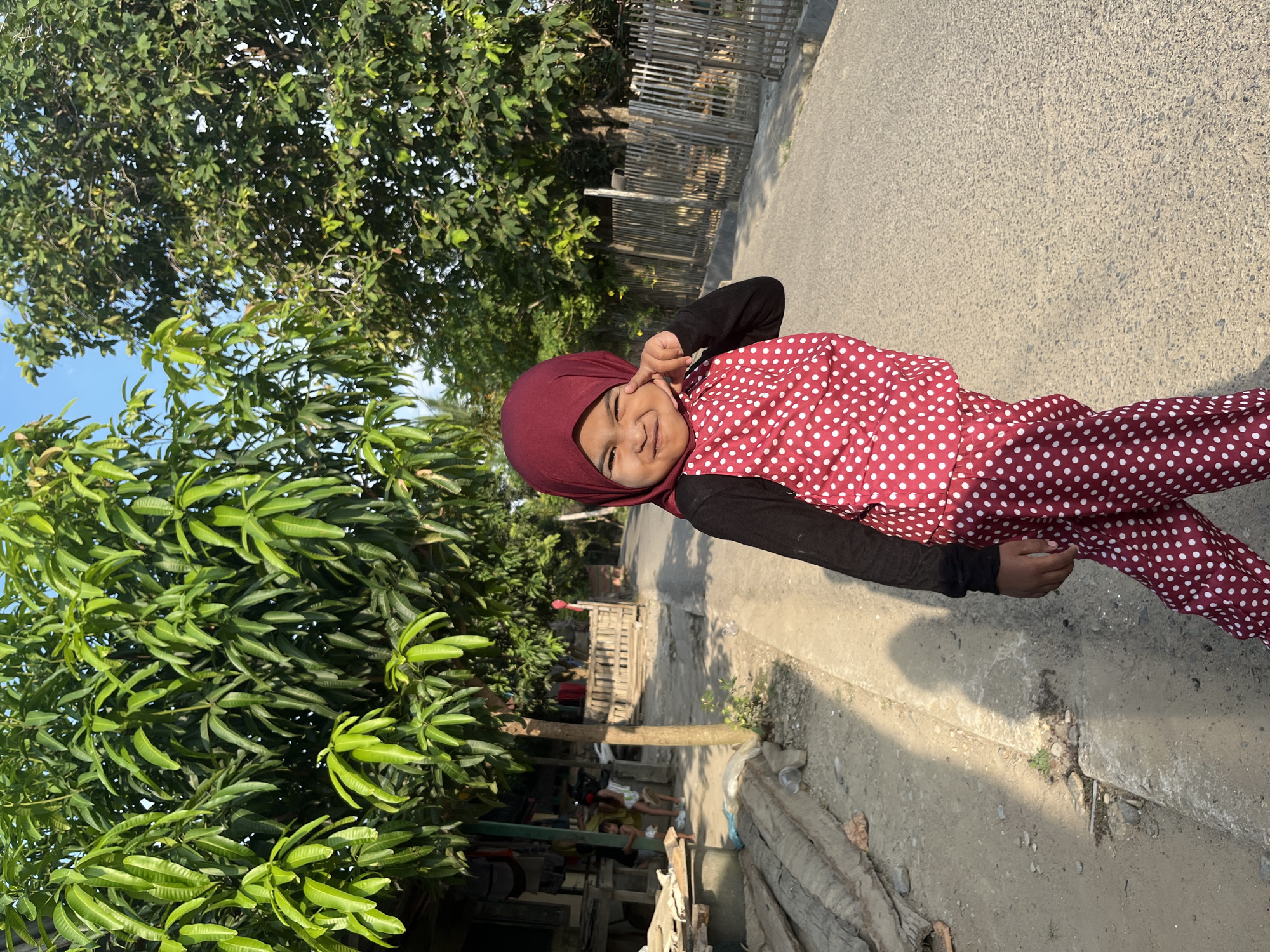 In Kruengmate, Indonesia, a young girl is captured in a full-body portrait outdoors. She wears a dark long-sleeved shirt, a red hijab, and matching red polka-dot pants and vest. The girl is positioned on the right side of the frame, facing the camera with her left hand raised to her face, and her right hand extended slightly forward. She has a playful expression, with her eyes squinted and a slight smile. Behind her, a paved road stretches into the background, bordered by a bamboo fence on the right. Lush green trees with large leaves are visible on both sides of the road, with one prominent tree to the left of the girl and another to the right. Sunlight casts shadows on the ground to the left, revealing a simple dirt path with some scattered debris and wooden planks. Further in the background, on the left, a modest dwelling with a wooden structure and laundry hanging can be observed, along with two other children playing. The sky is clear and blue.