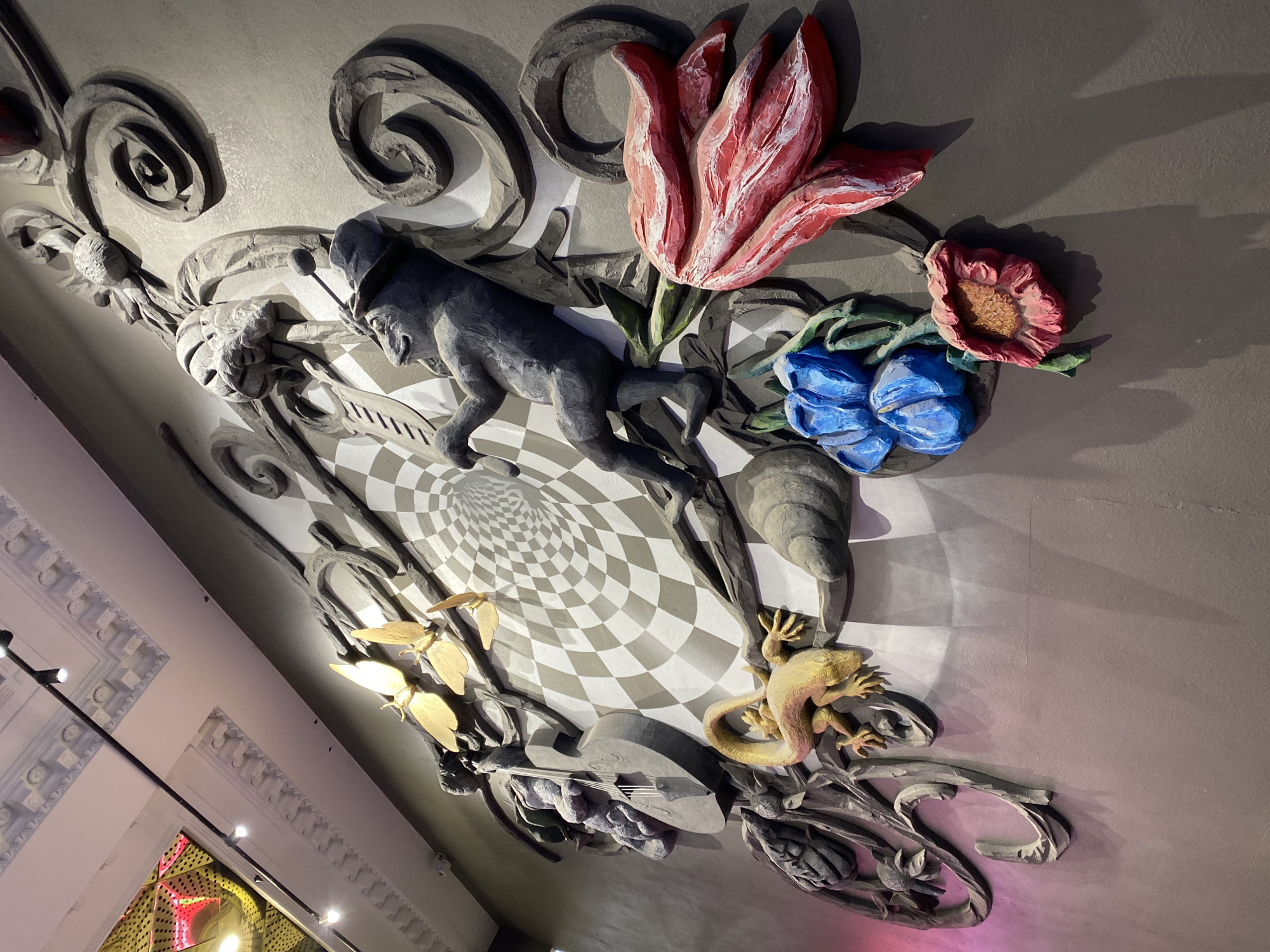 The image shows a ceiling mural inside the Weihsler Textile Factory building in Yekaterinburg, Russia. The mural is a three-dimensional installation featuring a variety of sculpted elements. These include flowers (a red and white tulip, a pink and orange flower, two blue flowers), a grey sculpted man, a lizard, butterflies, musical instruments (guitar), and swirls that appear to frame other features. The mural has a stylized checkerboard background, adding to the visual complexity. The mural is illuminated by an unknown source, which highlights the three-dimensional aspects. The scene's environment appears to be the interior of a building, with architectural details like ornate moldings visible along a wall to the left.