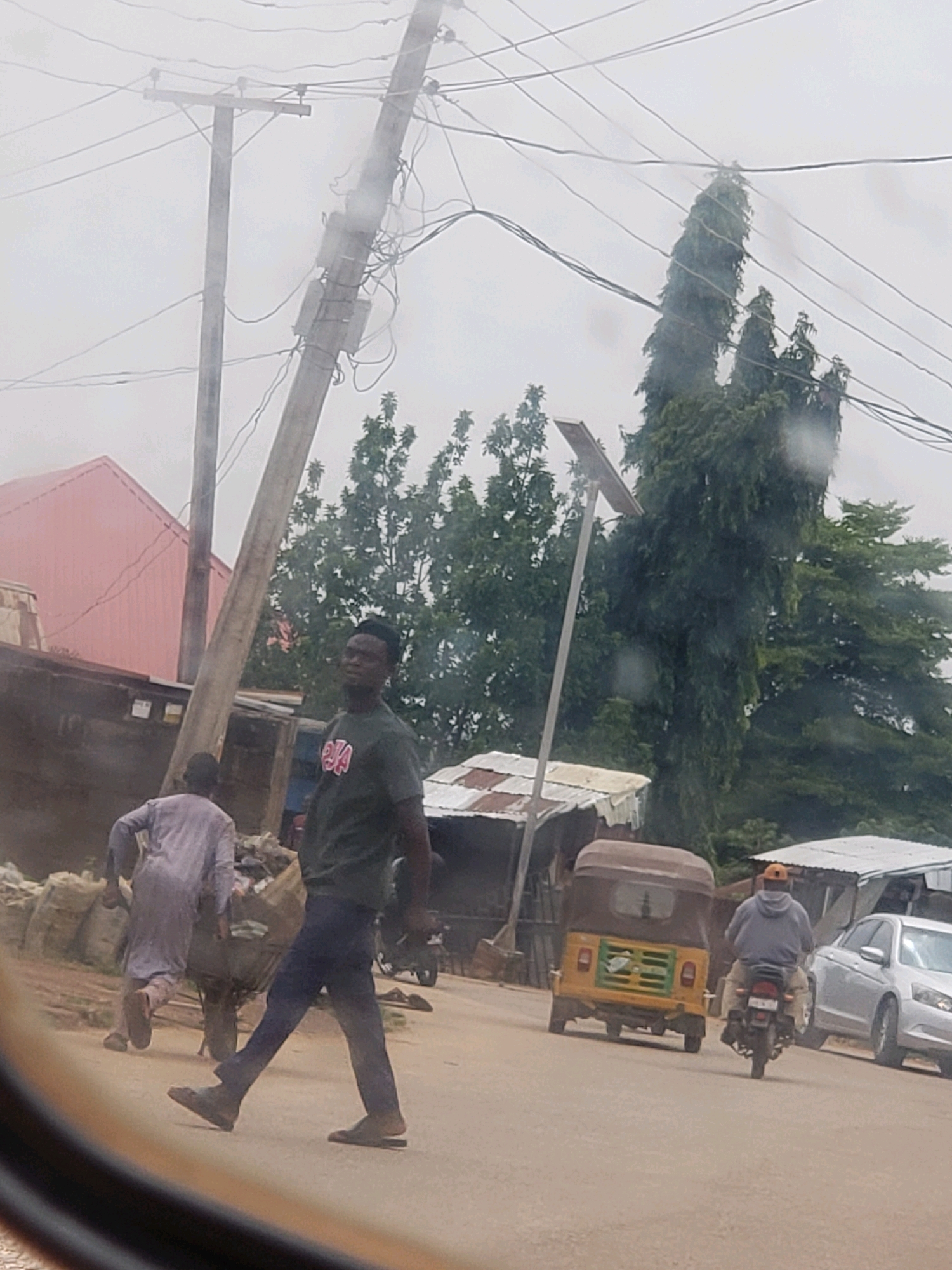 In Narayi, Nigeria, on a cloudy day, a person in a dark t-shirt and dark pants walks across a paved road. The person is looking to their left. Behind them, another person in a light-colored robe pushes a cart loaded with items. In the background, a yellow auto-rickshaw is parked on the side of the road, and a silver car is visible behind it. A person wearing a grey hooded sweatshirt and an orange cap rides a motorcycle away from the camera. Several telephone poles and wires are present in the scene, along with trees and some corrugated metal structures.