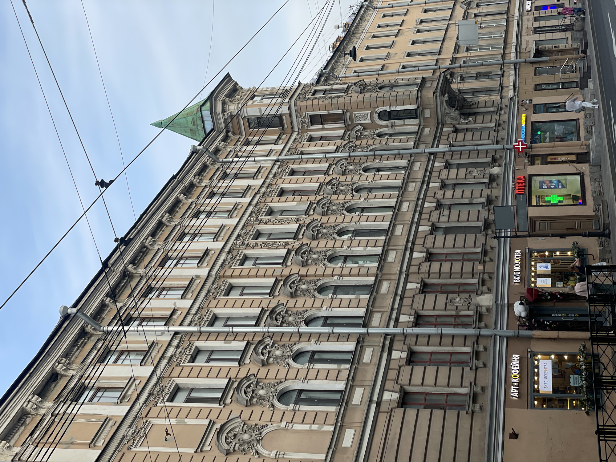 This image captures a street scene on Fontanka River Embankment, 38, in Saint Petersburg, Russia, featuring a multi-story, ornate historical building under an overcast sky. The building, constructed in an early 20th-century architectural style, displays a light beige facade with intricate classical revival decorative elements, including arched window surrounds, pilasters, and cornices. A prominent corner section of the building features a tall, octagonal turret topped with a green, conical spire. Multiple dark-framed windows are visible across the building's facade, with some on the second story exhibiting arched tops. Several grey metallic downspouts run vertically along the building.
The ground floor houses several commercial establishments. From left to right, visible businesses include "АРТ+КОФЕЙНЯ" (ART+COFFEE SHOP) and "ВКУС ИСКУССТВА" (TASTE OF ART), both featuring illuminated signs, large display windows, and decorative elements such as flower boxes and a white cloud-shaped sculpture above the shared entrance. Further to the right is an "АПТЕКА" (PHARMACY), identifiable by its red sign and a prominent green cross symbol, with a separate green LED cross on its window. Beyond the pharmacy, additional commercial storefronts are partially visible.
Utility lines, likely for trolleybuses or other utilities, crisscross the upper portion of the frame against the light blue and grey sky. On the sidewalk in front of the shops, several individuals are present. Two people are near the entrance of "ВКУС ИСКУССТВА." Another person, dressed in light clothing, walks along the sidewalk in front of the "АПТЕКА." Further right, two more individuals, one in a pink garment and another with a small dog, are also walking. A black metal railing separates the sidewalk from the asphalt street in the foreground. The overall scene depicts a typical urban environment in Saint Petersburg, characterized by historic architecture and active commercial spaces.