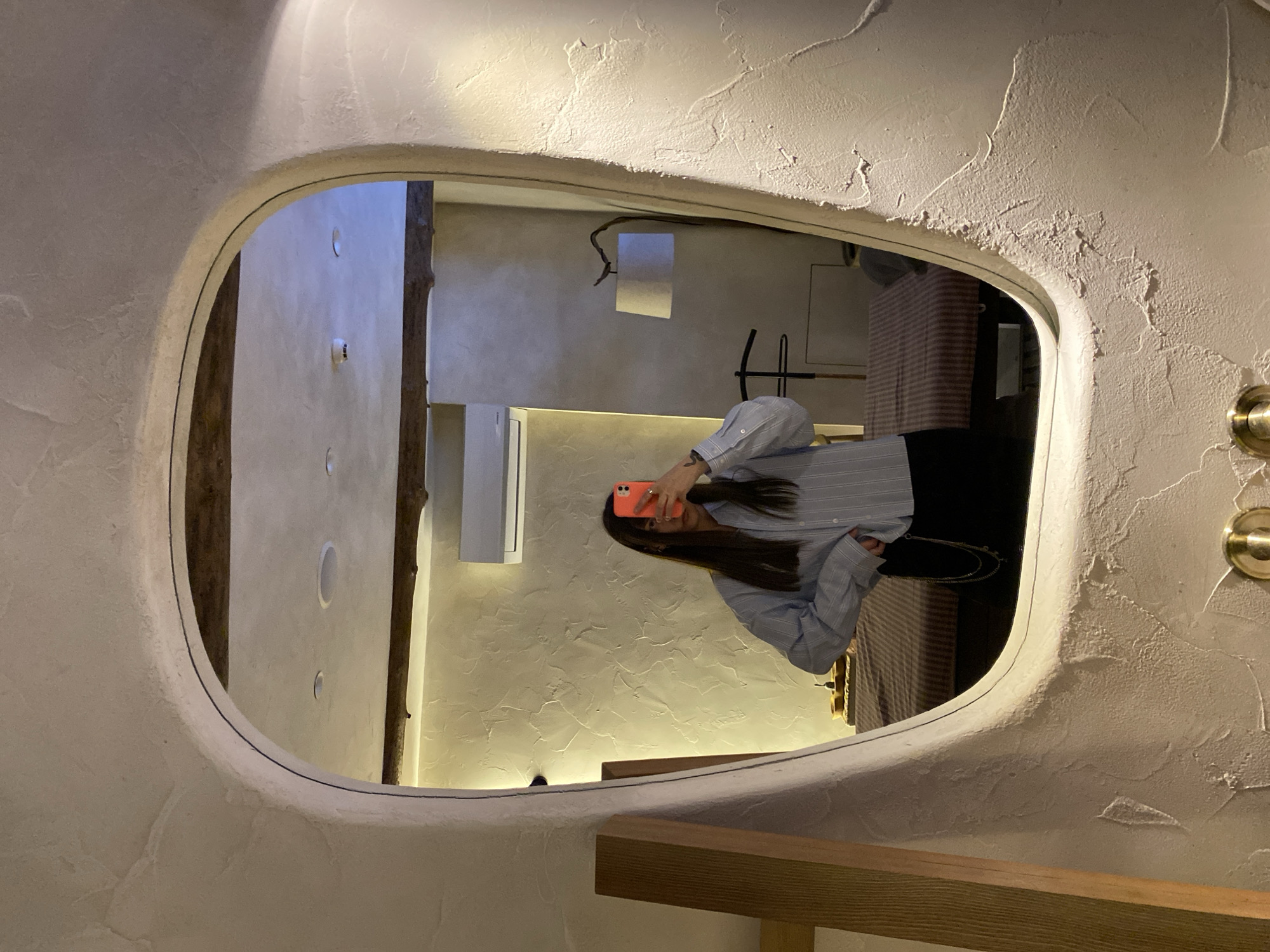 A reflection of a woman taking a photo in a uniquely framed mirror. The mirror is surrounded by a white, textured wall. The reflection shows interior architecture with white walls and what appears to be a wooden support beam. The woman is holding an orange cellphone and wearing a light blue striped shirt. The reflection also captures a striped object in the background.